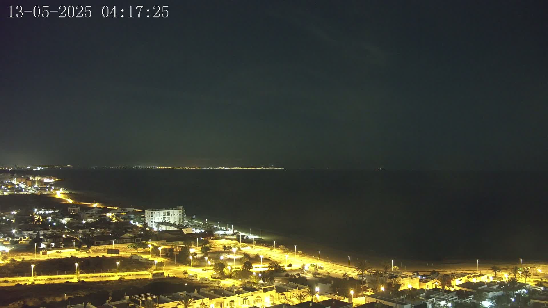 A nighttime coastal town with illuminated streets and buildings is situated next to a dark sea under a clear, dark sky.