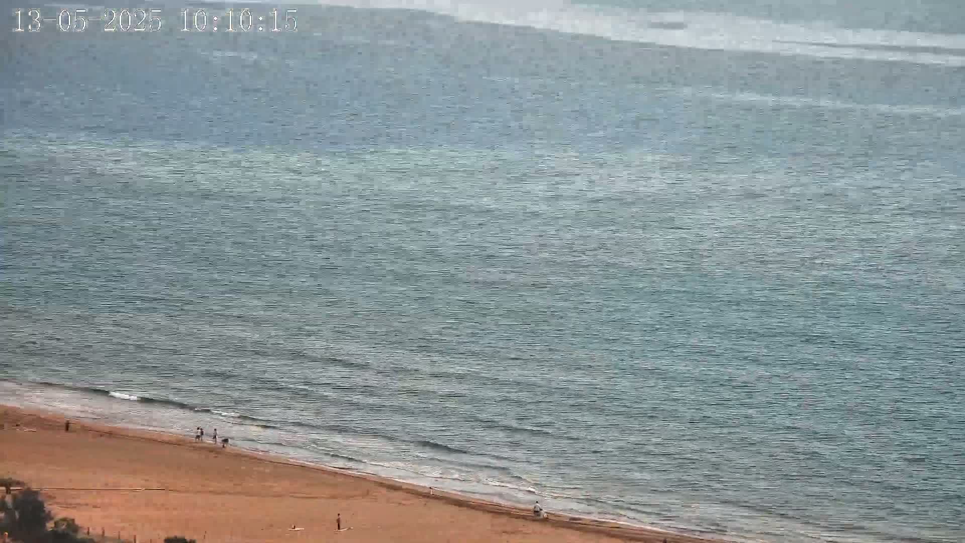A sandy beach with a few people on it slopes down to a calm, teal sea under a mostly clear sky.
