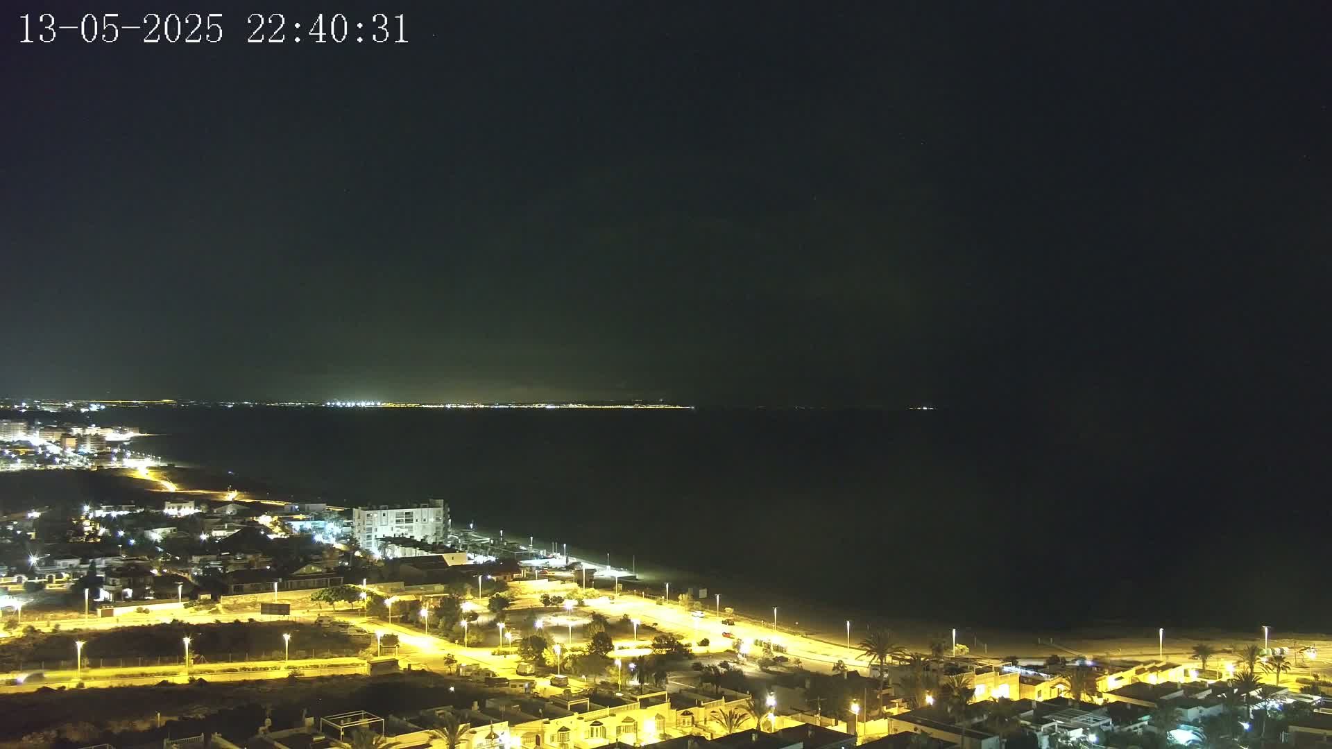 A nighttime view shows a dark ocean with a distant, faintly lit coastline, and a closer, brightly lit coastal town with roads and buildings.