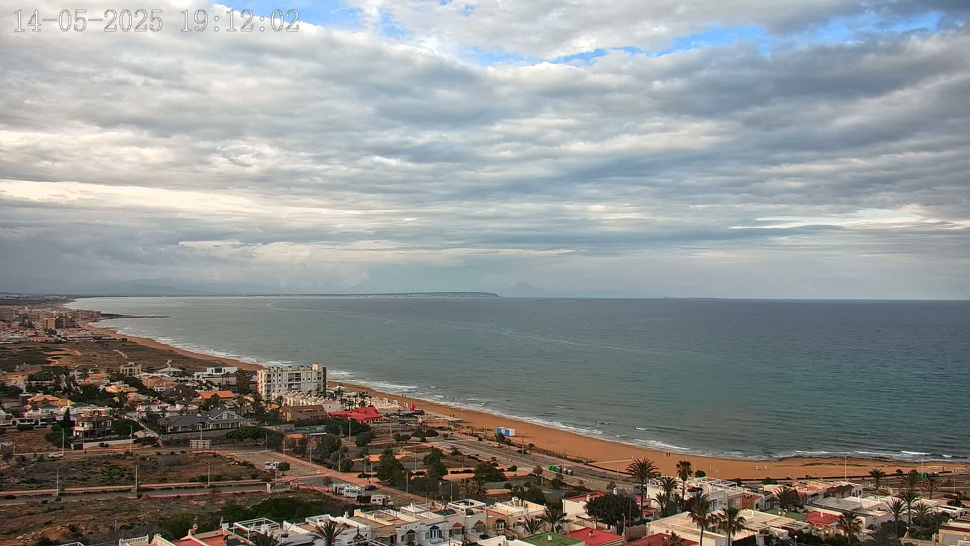 A coastal town with low-rise buildings is situated next to a sandy beach and a calm sea under a mostly cloudy sky.