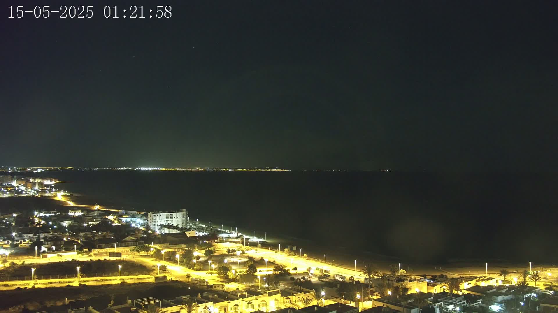 A nighttime coastal scene shows a dark sea under a clear sky, with a brightly lit town situated along the beach.