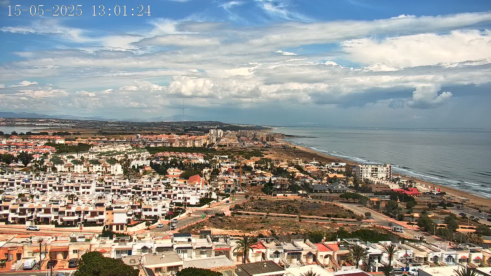 A coastal town with numerous white buildings stretches along a sandy beach under a partly cloudy sky.