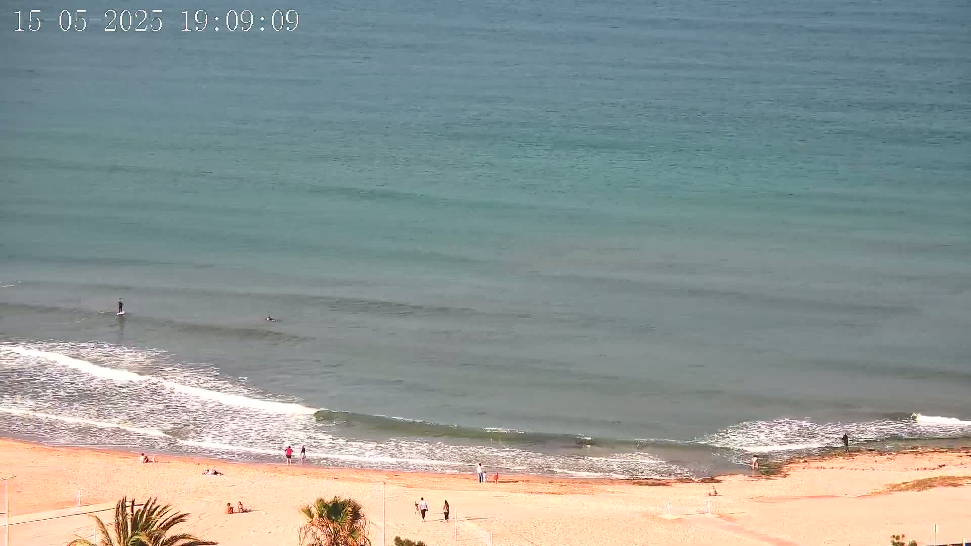 A sandy beach with a few people on it is met by a calm, light-blue ocean with small waves.