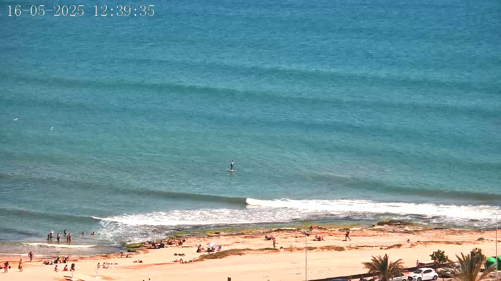 An overhead view shows a sandy beach with several people sunbathing and swimming in the ocean, where a person is paddleboarding under a sunny sky.
