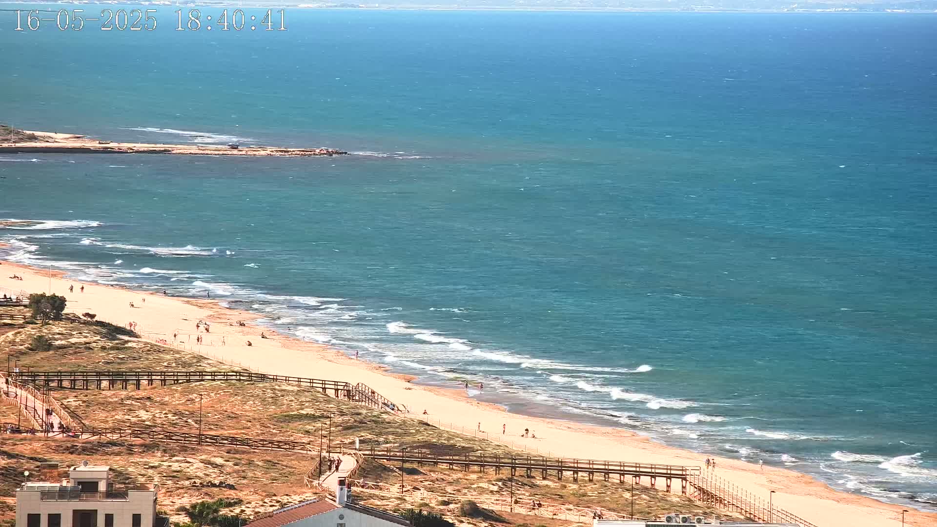 A sandy beach with a few people on it, bordered by a wooden walkway and dunes, meets a calm, teal ocean under a sunny sky.