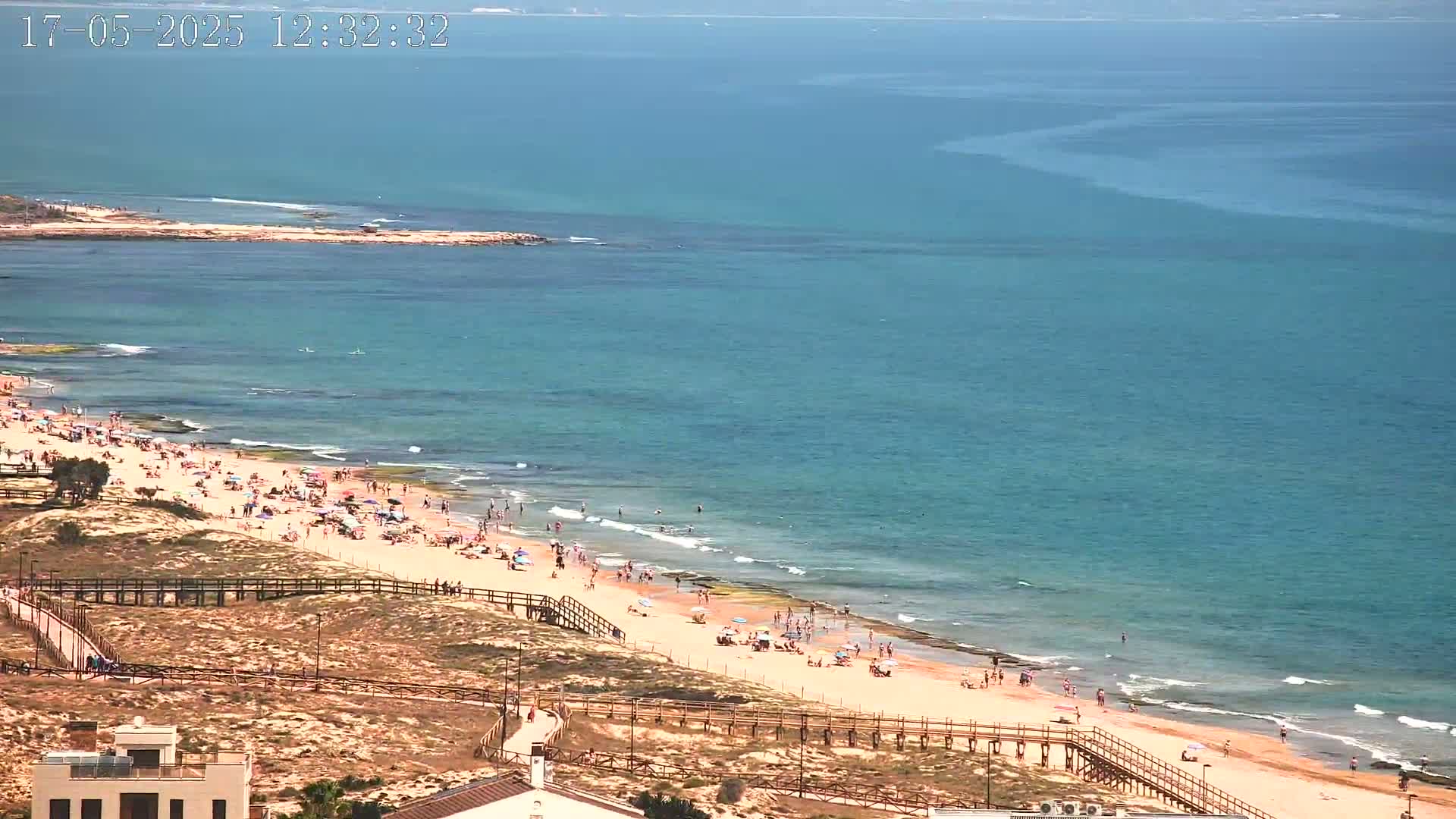 A sunny day at a sandy beach with many people enjoying the water and sun, viewed from an elevated position.
