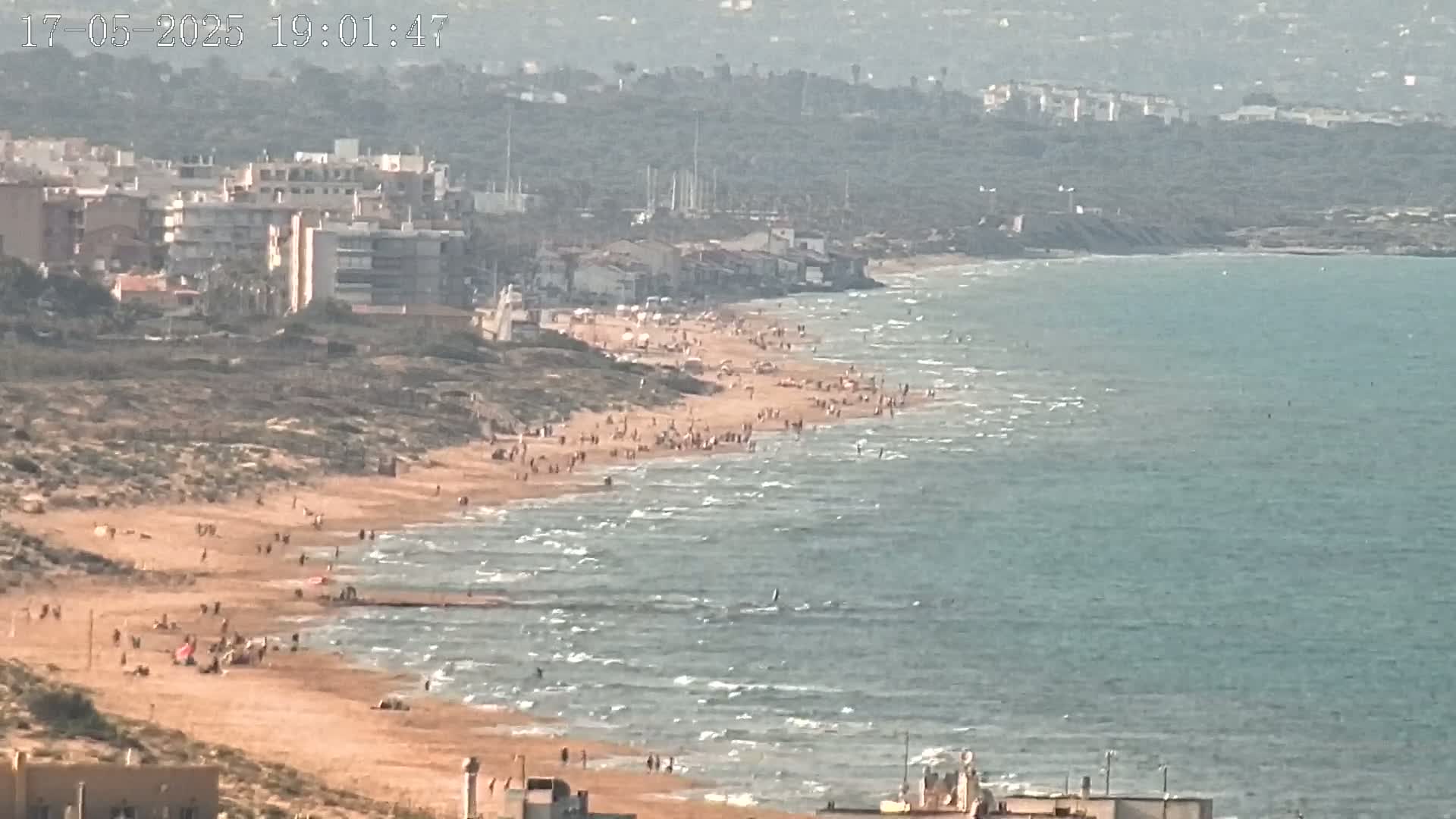 A sandy beach with many people on it slopes gently into a calm, light-blue sea near a town of low-rise buildings under a hazy sky.