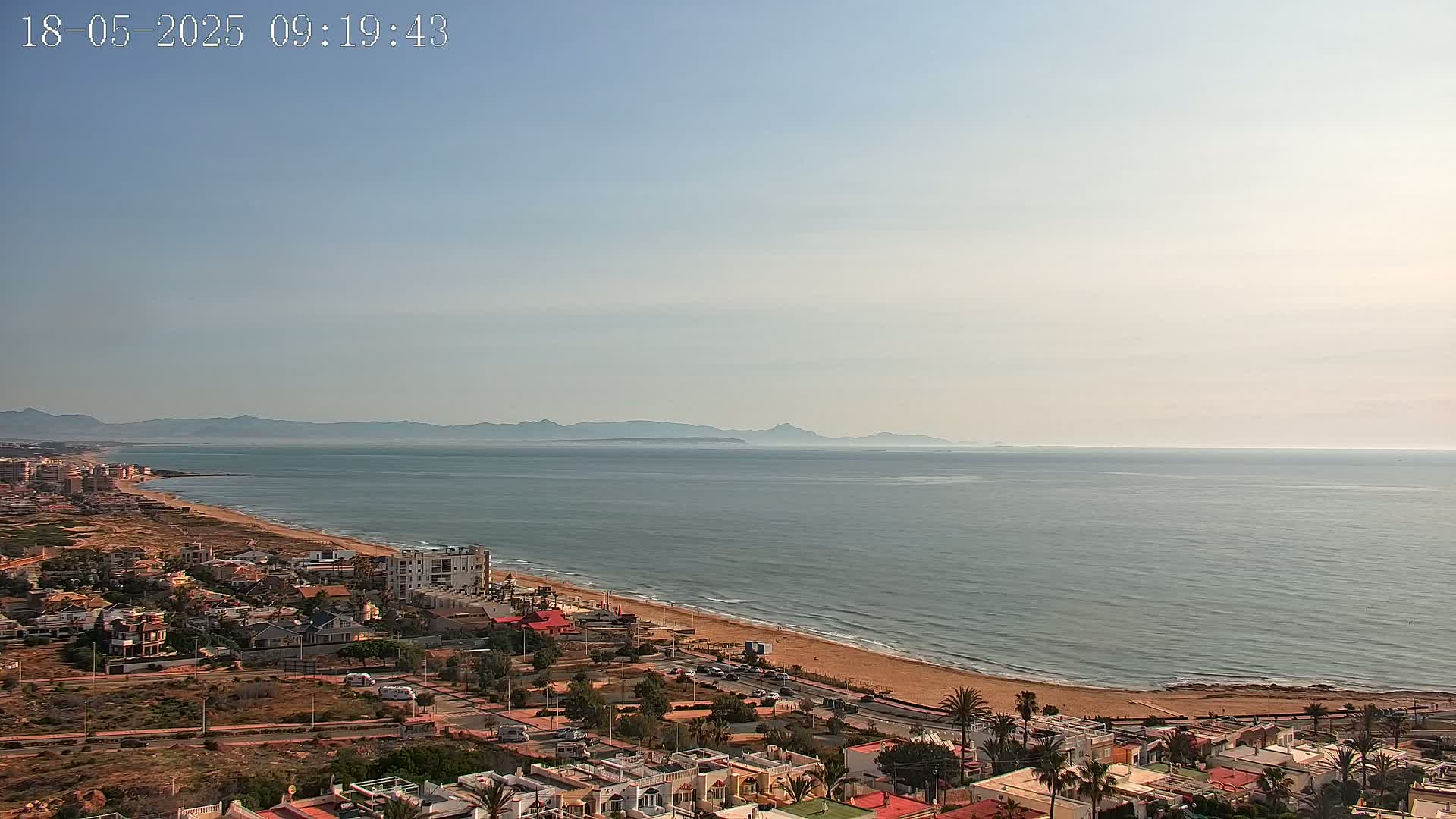 A coastal town overlooks a calm sea and a distant mountain range under a clear sky.