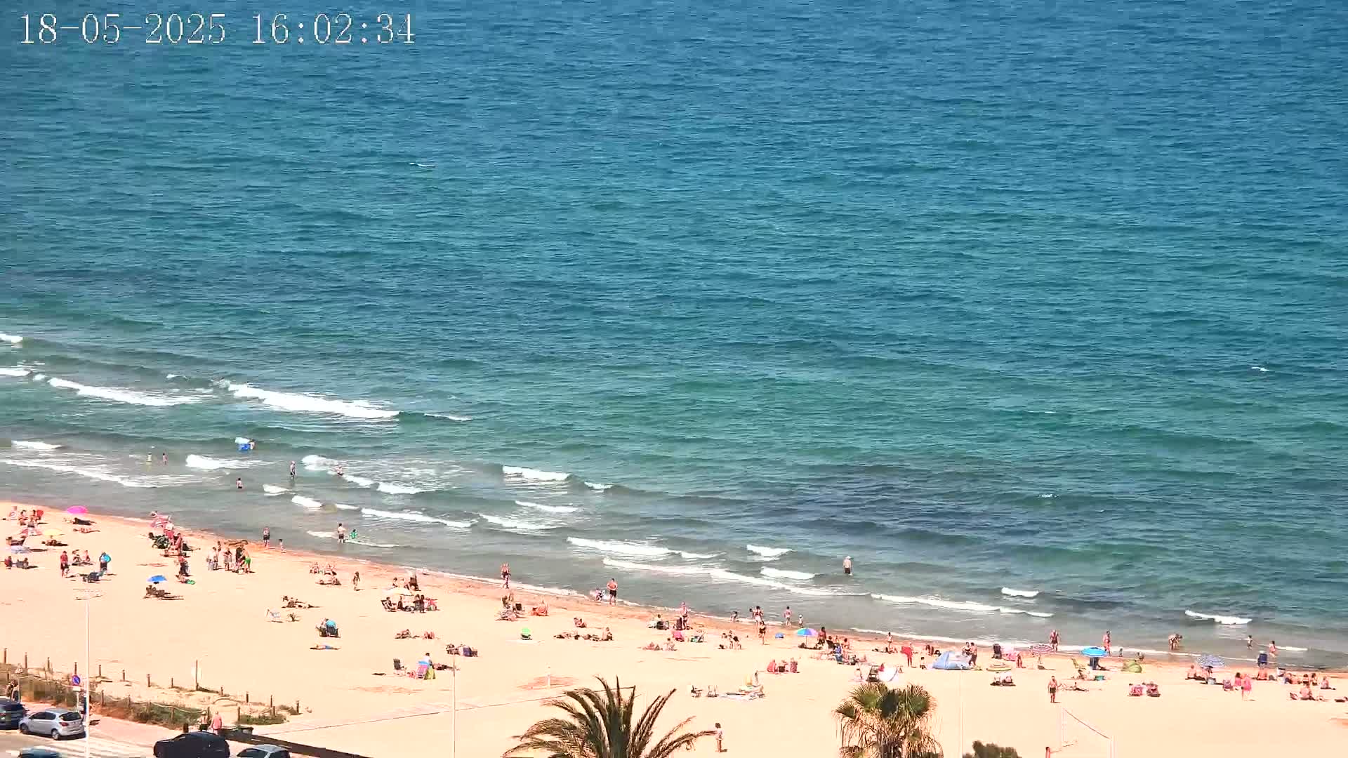 A sandy beach with many people sunbathing and swimming in a calm, blue ocean under a sunny sky.