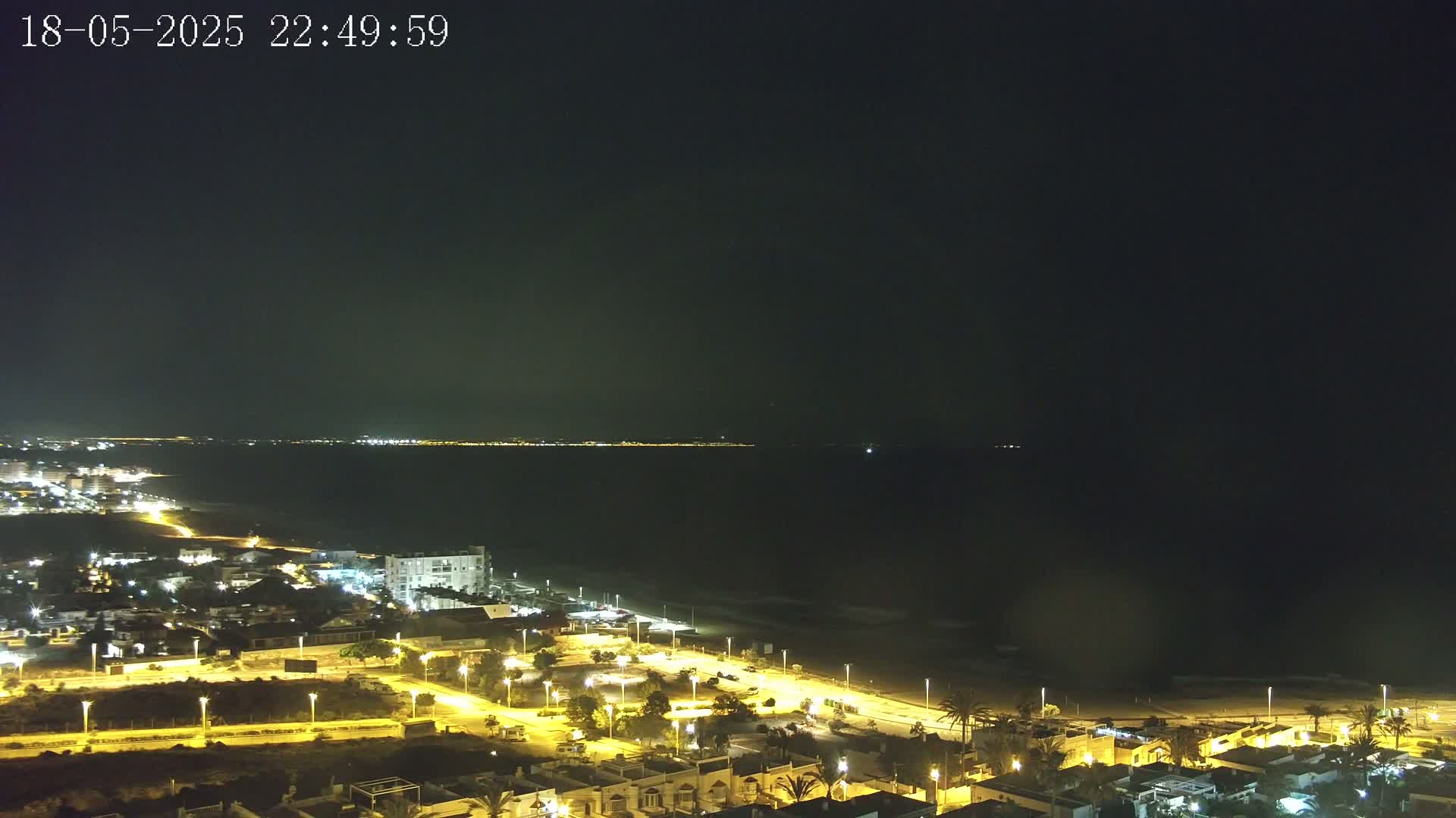A night-time view of a coastal town illuminated by streetlights, overlooking a dark ocean with a faintly visible distant shoreline.