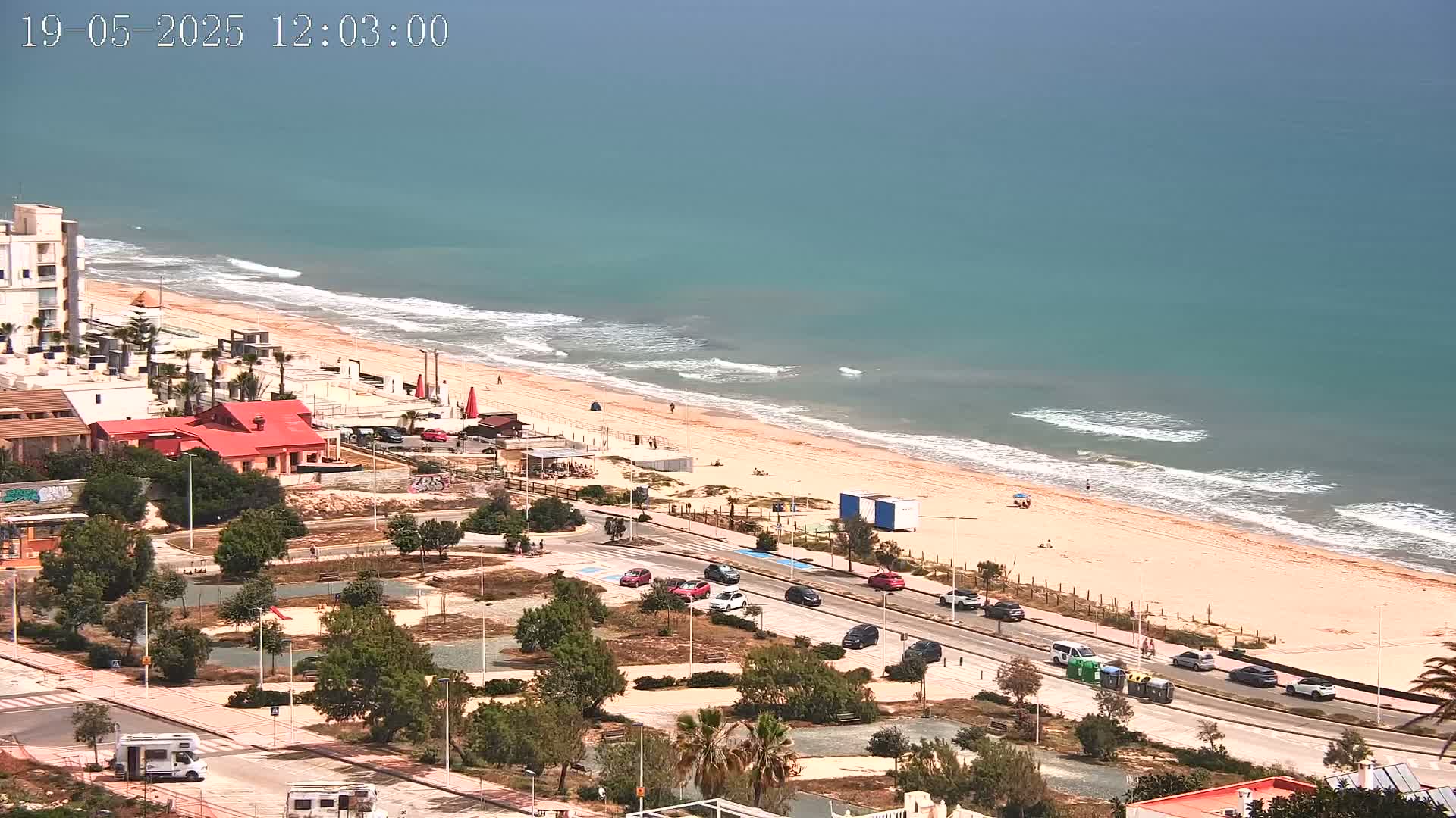 An aerial view shows a sandy beach with gentle waves under a hazy sky, a road with cars parked along it, and buildings and vegetation behind it.