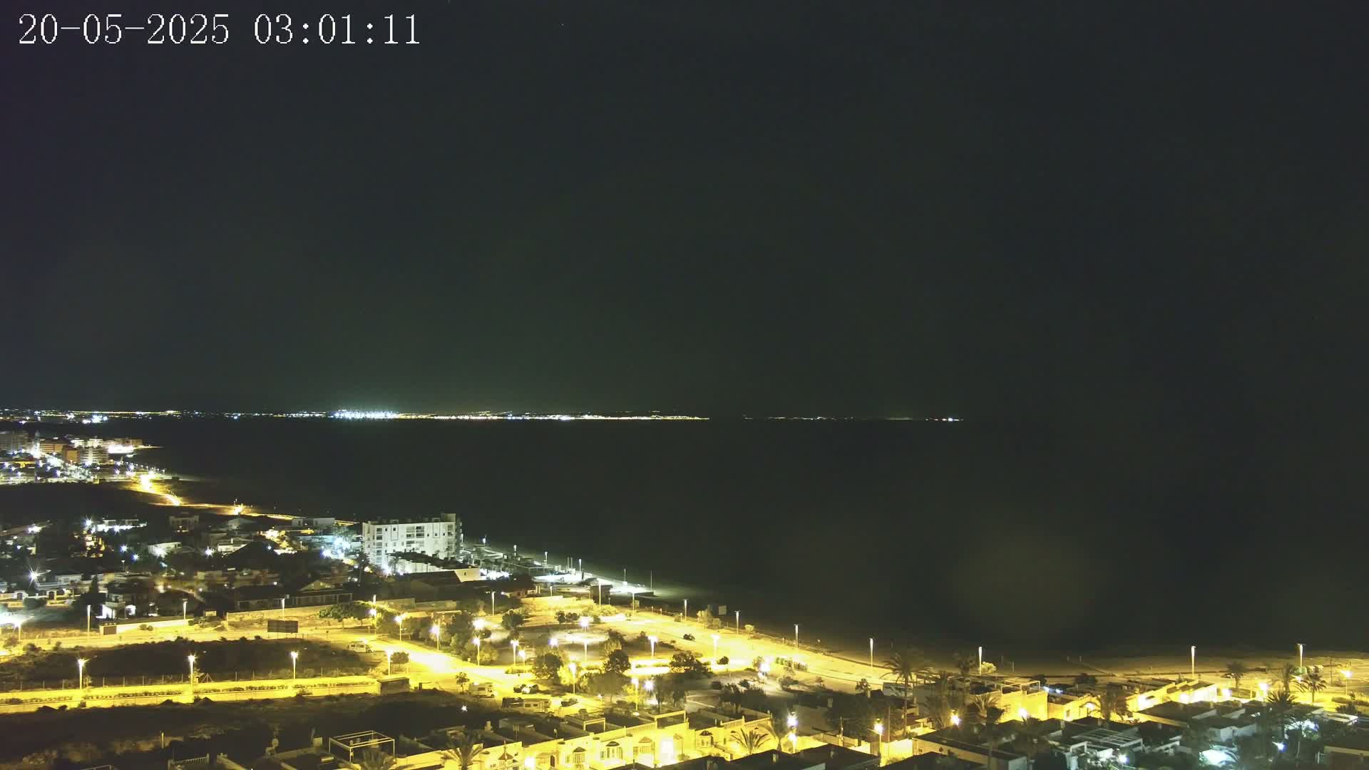 A nighttime coastal scene shows a dark body of water with a faintly lit distant shoreline, and a closer, brightly lit residential area stretching towards the beach.