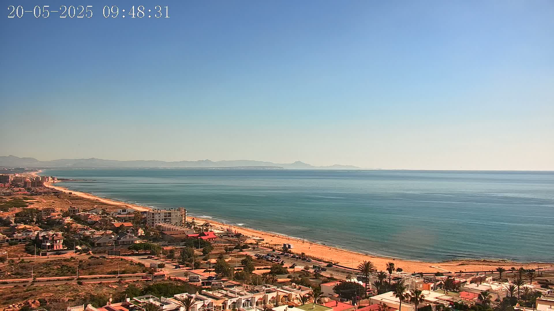 A clear, sunny day reveals a coastal town overlooking a long sandy beach and calm blue ocean, with distant mountains visible on the horizon.