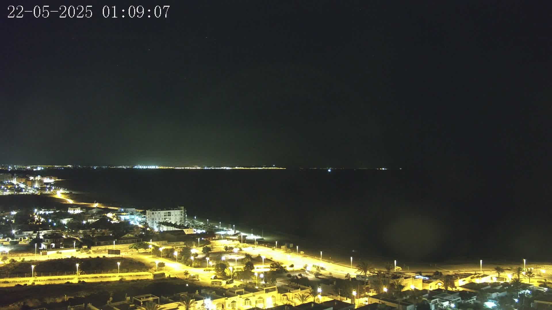 A nighttime coastal town is illuminated by streetlights, overlooking a dark body of water with a distant city skyline faintly visible under a clear, starlit sky.