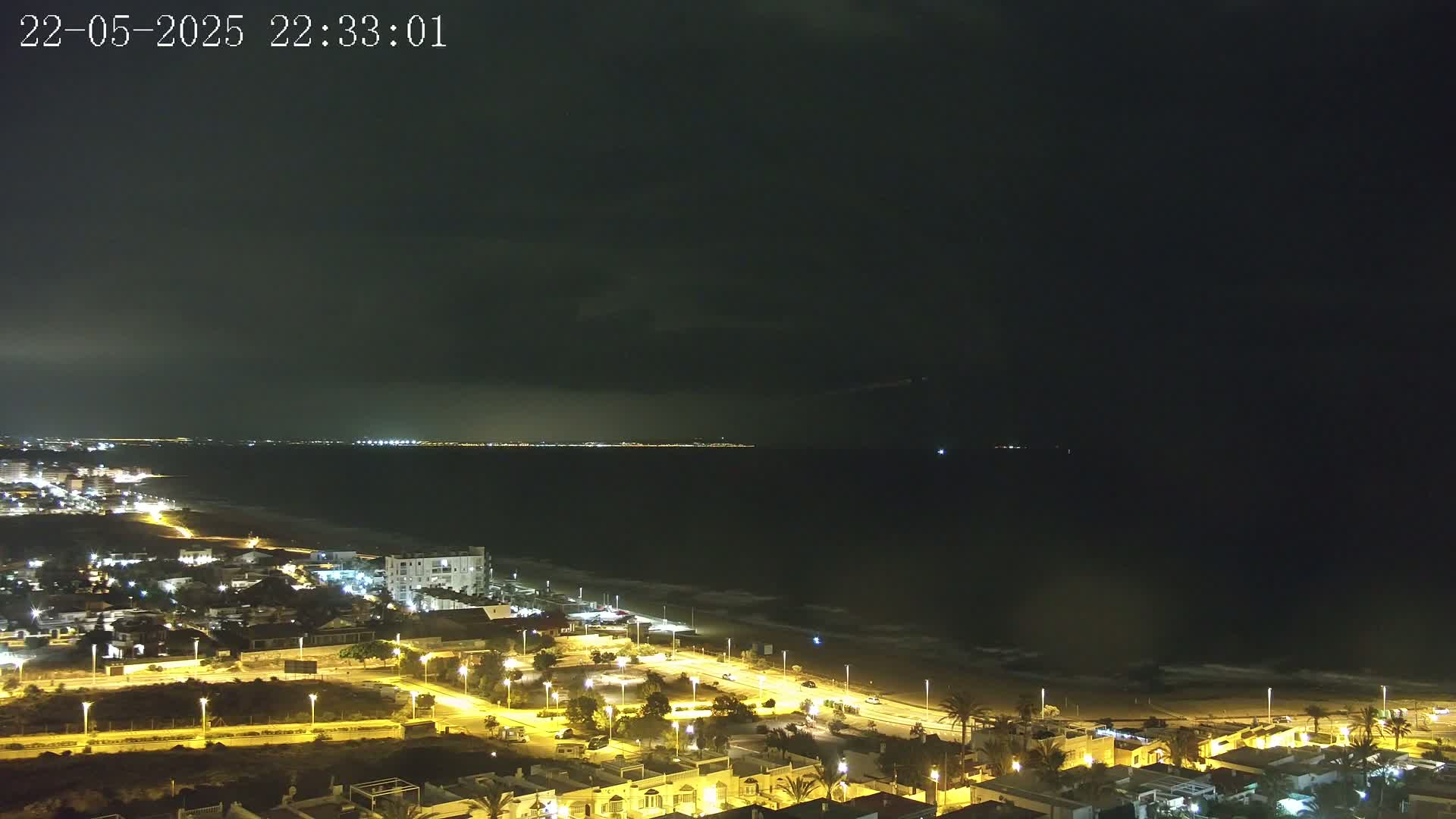A nighttime coastal scene shows a dark sea under a mostly cloudy sky, with a brightly lit beach and town visible in the foreground.