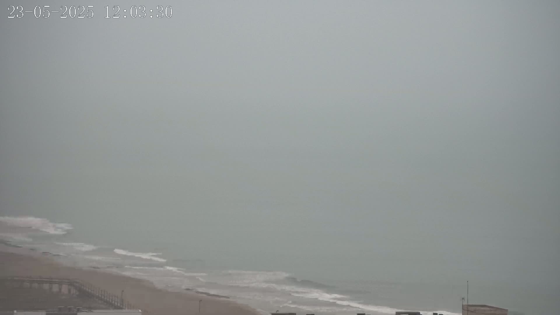 A hazy, overcast day shows a beach with gentle waves lapping the shore, and a pier partially visible in the foreground.