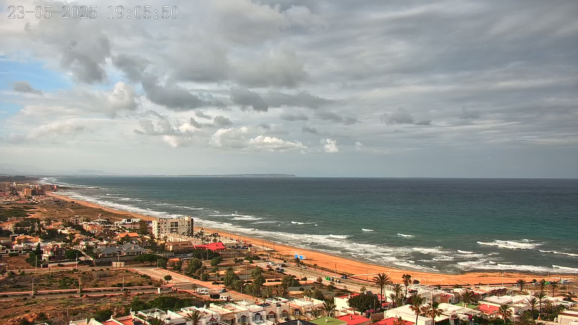 An expansive sandy beach with moderate waves stretches along a teal ocean under a partly cloudy sky.