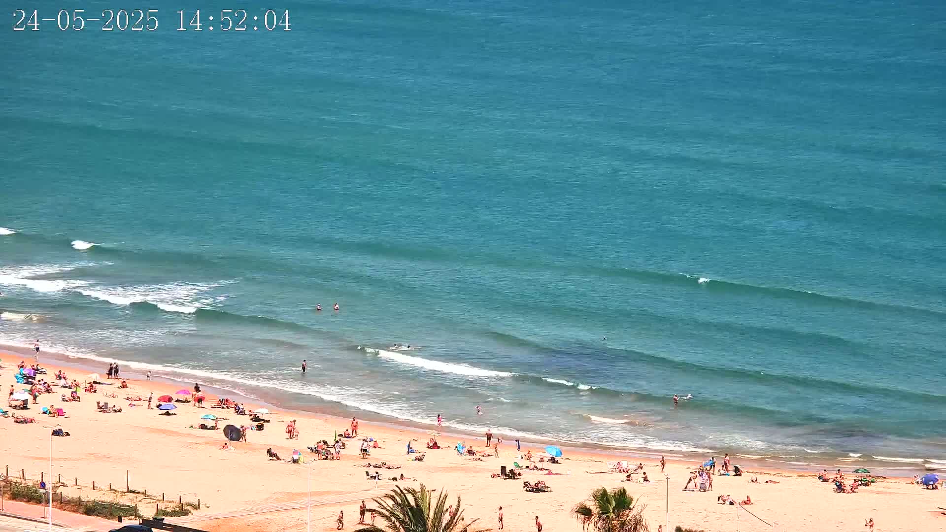 A sandy beach with many people sunbathing and swimming, bordered by a calm, turquoise ocean with gentle waves.