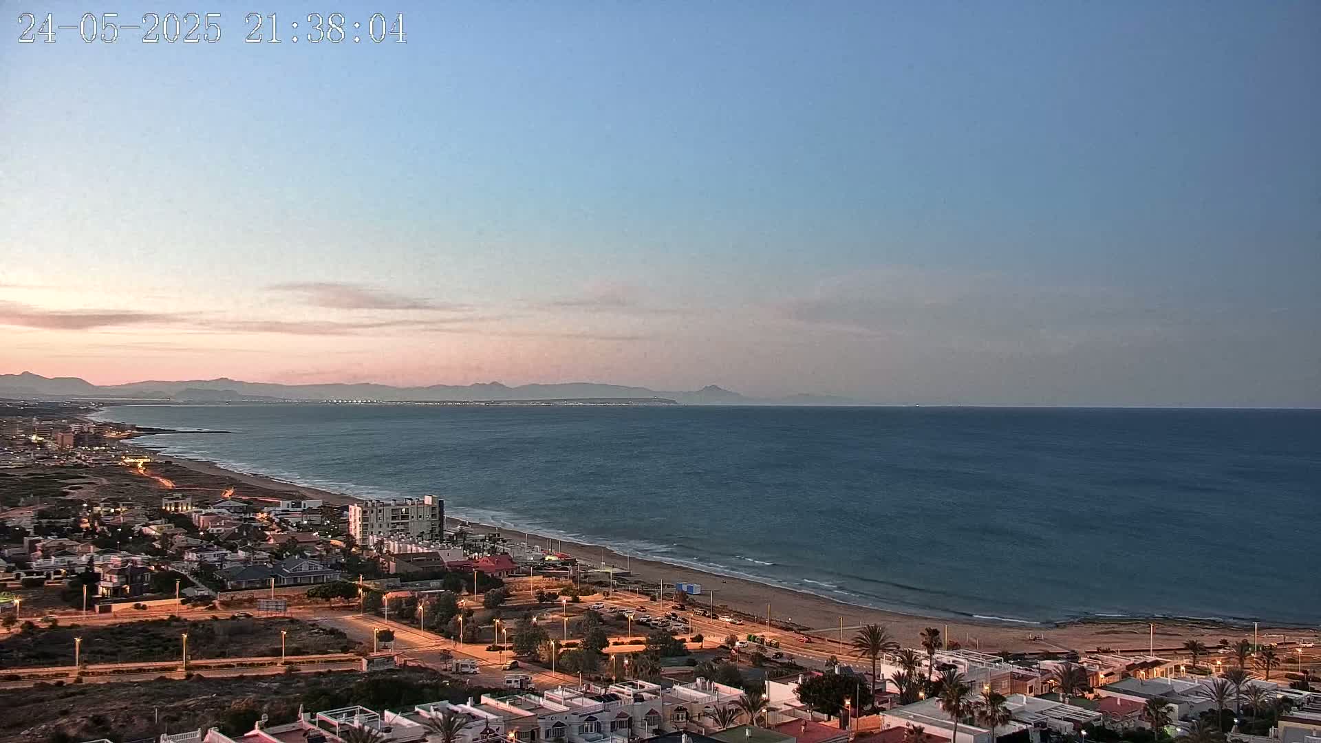 An aerial view shows a coastal town at dusk, with a calm ocean extending to distant mountains under a pale pink and blue sky.