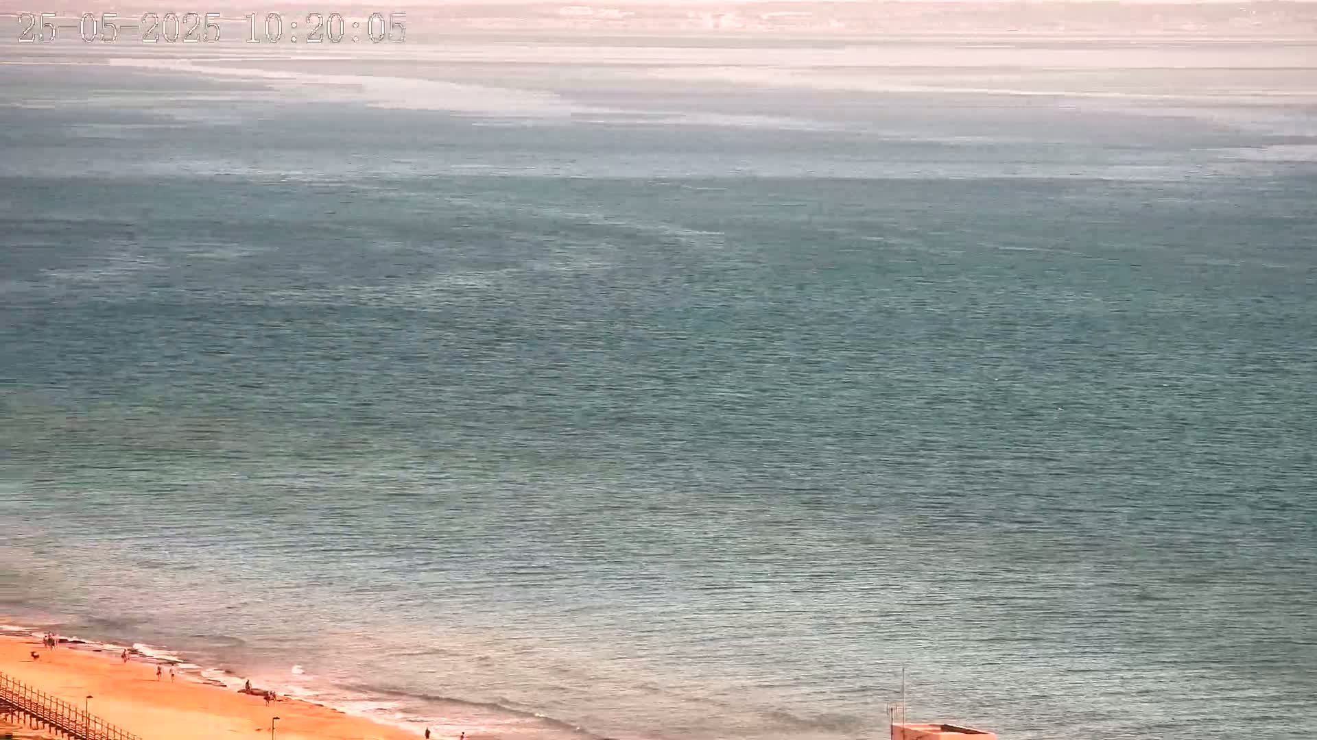 A calm, teal ocean meets a sandy beach with a few people visible on a sunny day.