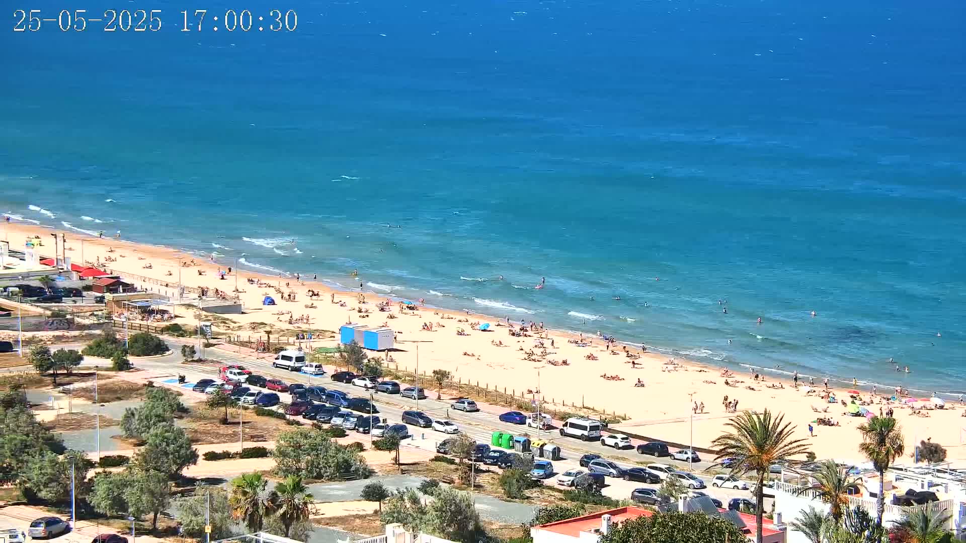A sandy beach crowded with sunbathers and swimmers borders a calm, turquoise ocean on a sunny day.
