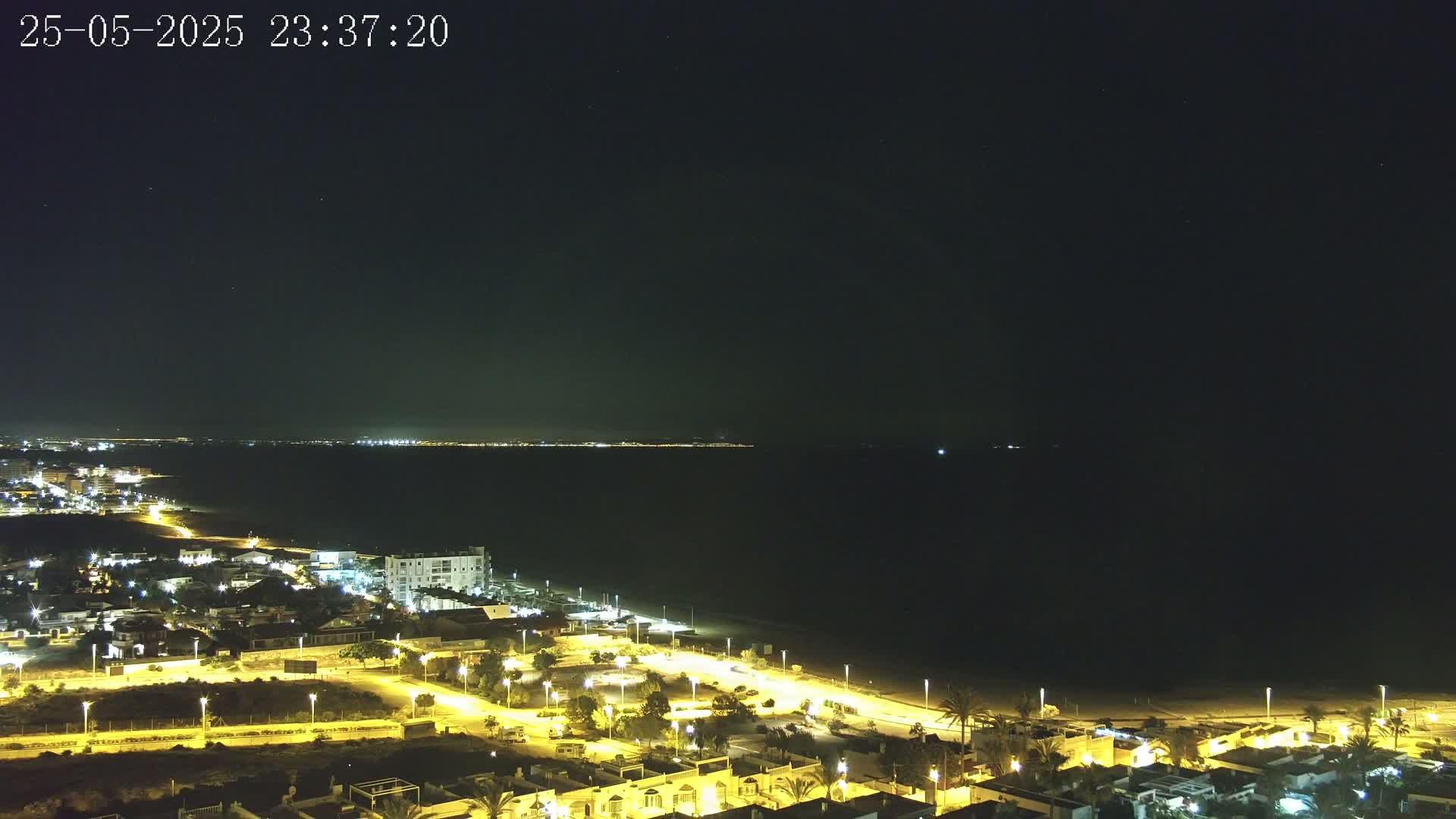 A nighttime coastal scene shows a dark body of water with a faintly lit distant shore, a beach, and a town with illuminated streets and buildings under a clear, starry sky.