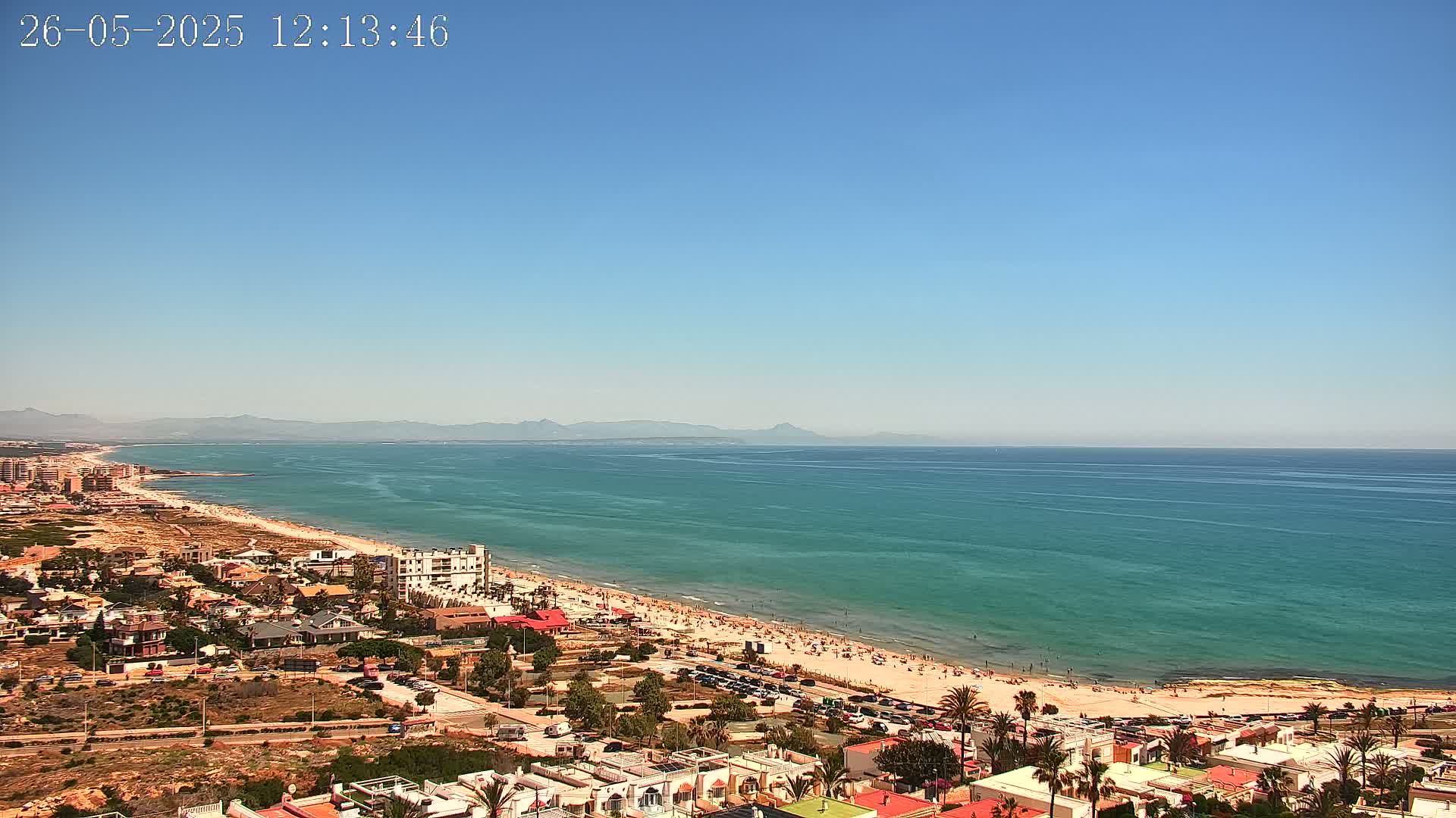 A clear, sunny day shows a long sandy beach crowded with people, adjacent to a town of low-rise buildings, all overlooking a calm, turquoise ocean with distant mountains.