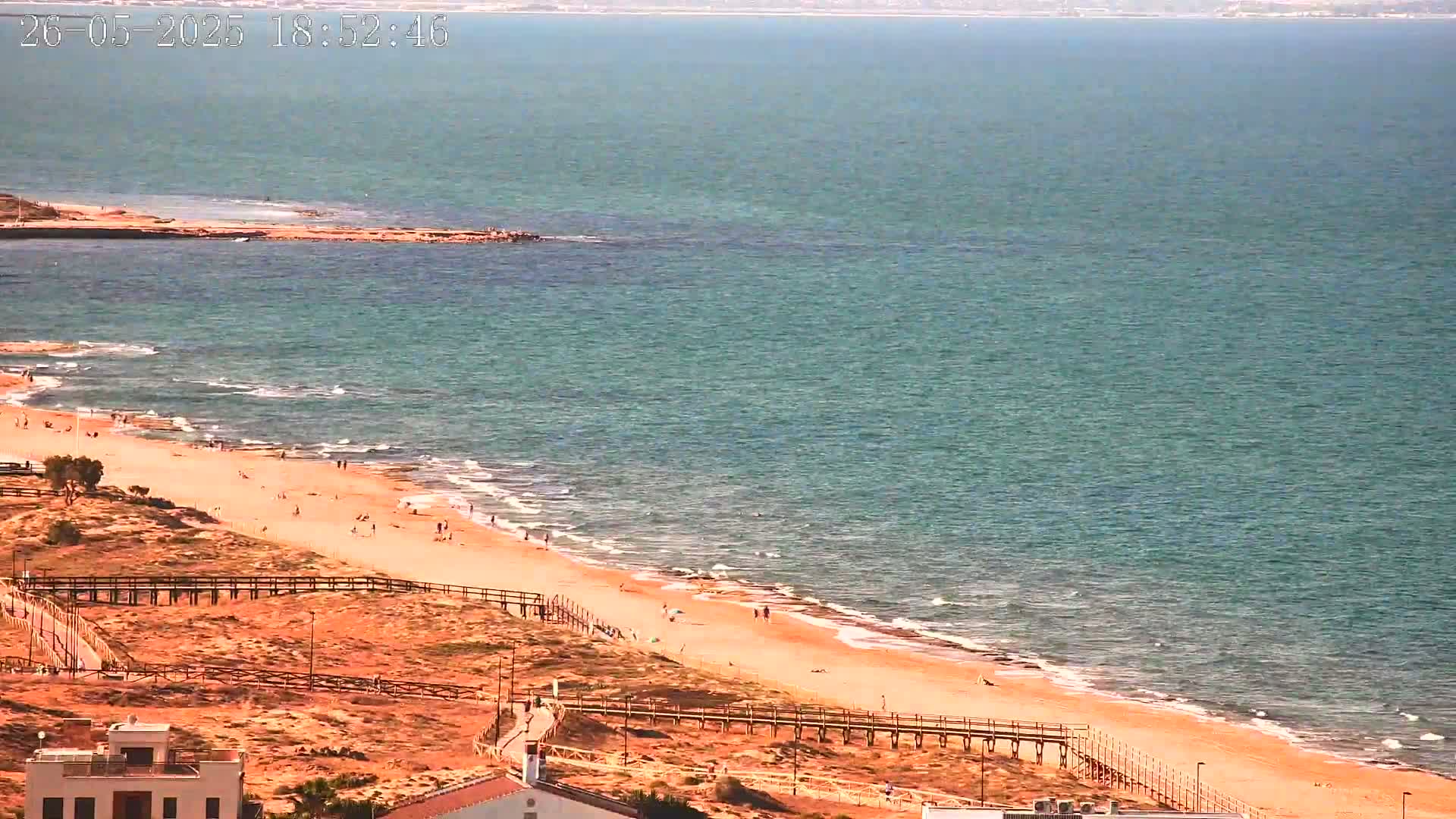 A sandy beach with a few people on it is bordered by a wooden walkway and overlooks a calm, teal ocean under a sunny sky.