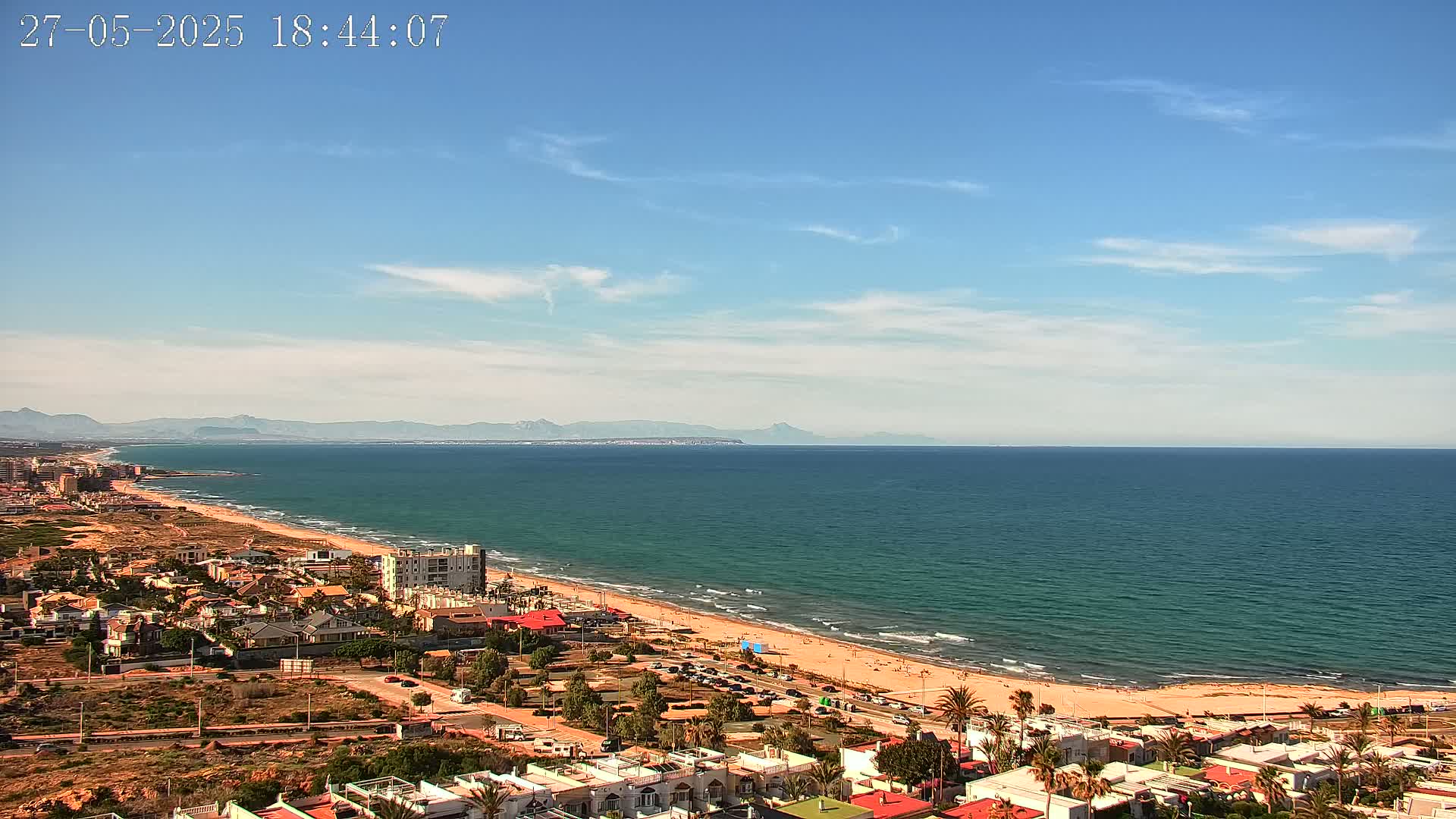 A coastal town with sandy beach stretches along a calm ocean under a mostly clear, sunny sky, with distant mountains visible on the horizon.