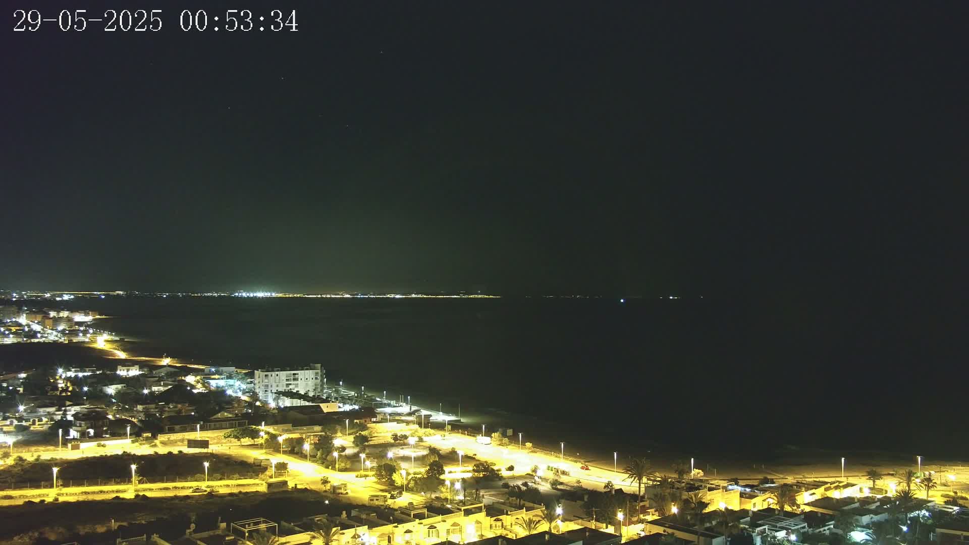 A nighttime coastal town with illuminated streets and houses overlooks a dark sea under a clear sky.