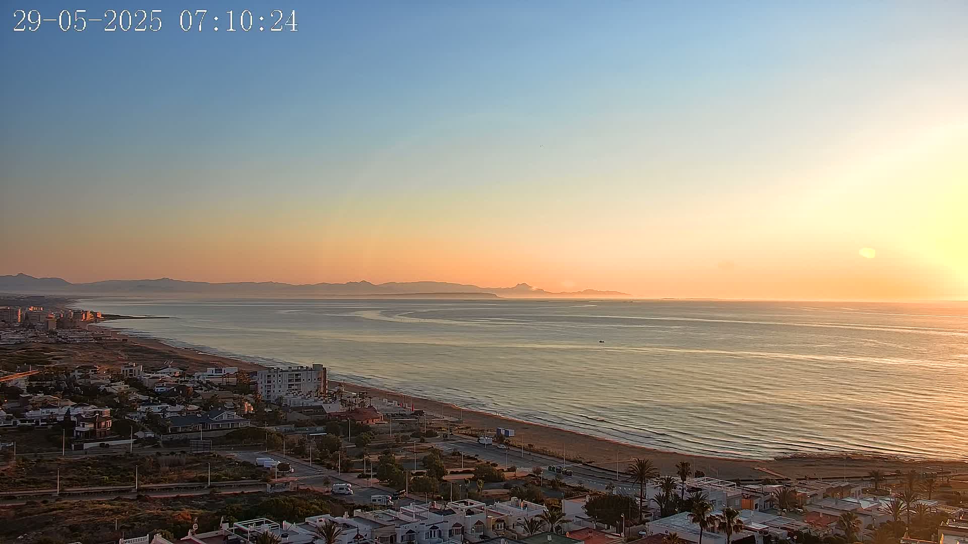 A coastal town at sunrise, with a calm ocean extending to distant mountains under a clear, orange-hued sky.