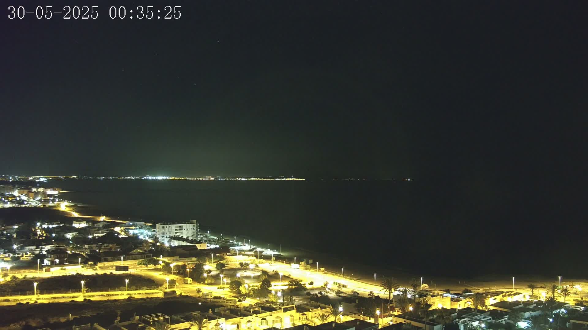 A nighttime coastal town with illuminated streets and buildings overlooks a dark body of water, under a clear, starry sky.