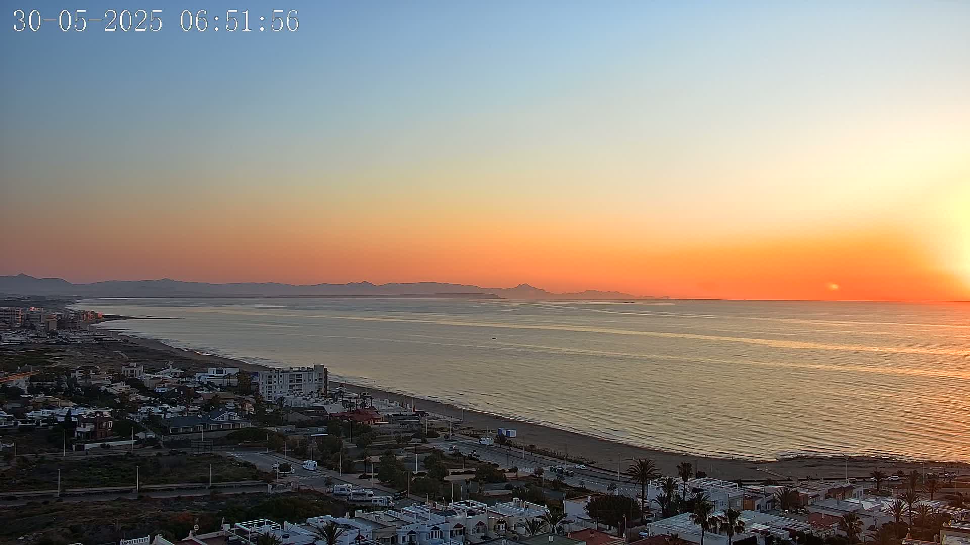 A coastal town at sunrise, with a calm sea reflecting the warm, orange hues of the sky and distant mountains on the horizon.