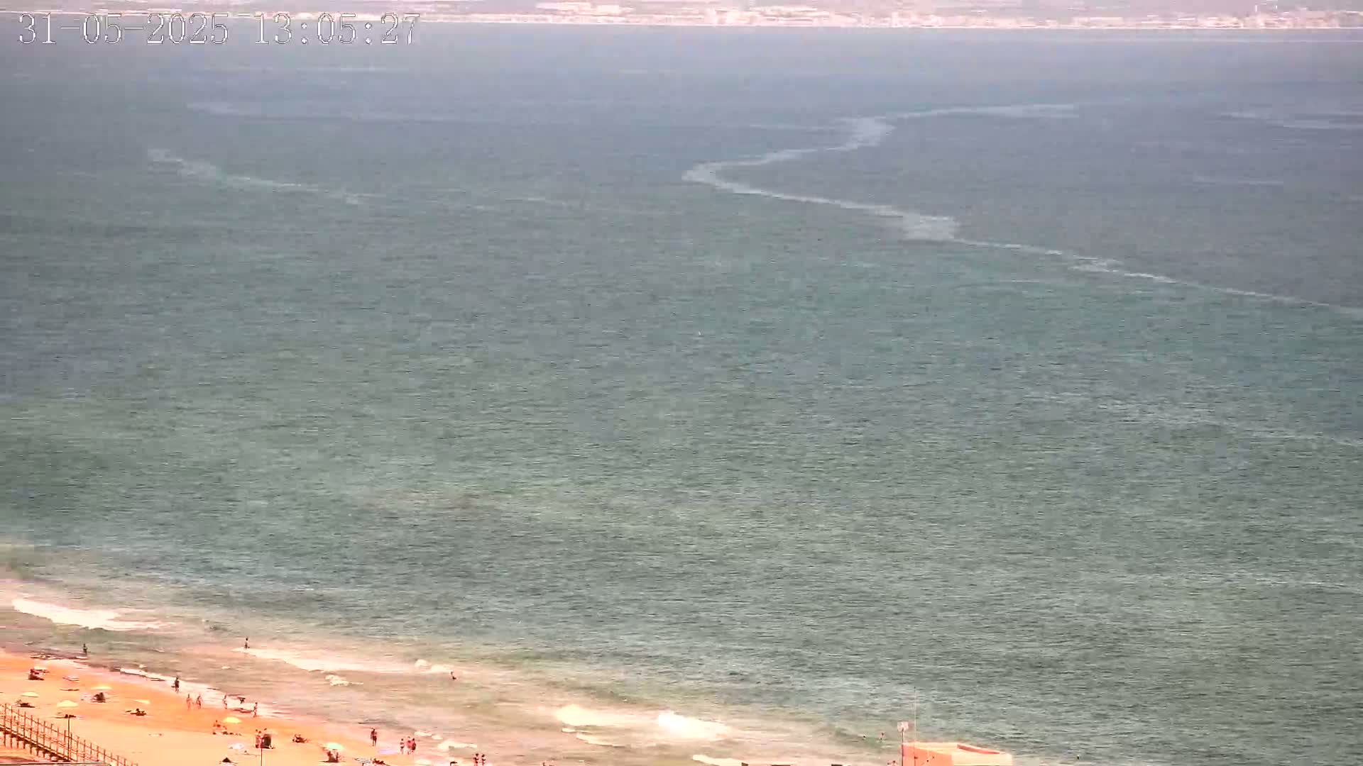A mostly sunny day shows a wide expanse of calm, greenish-blue ocean meeting a sandy beach with scattered people enjoying the shore.