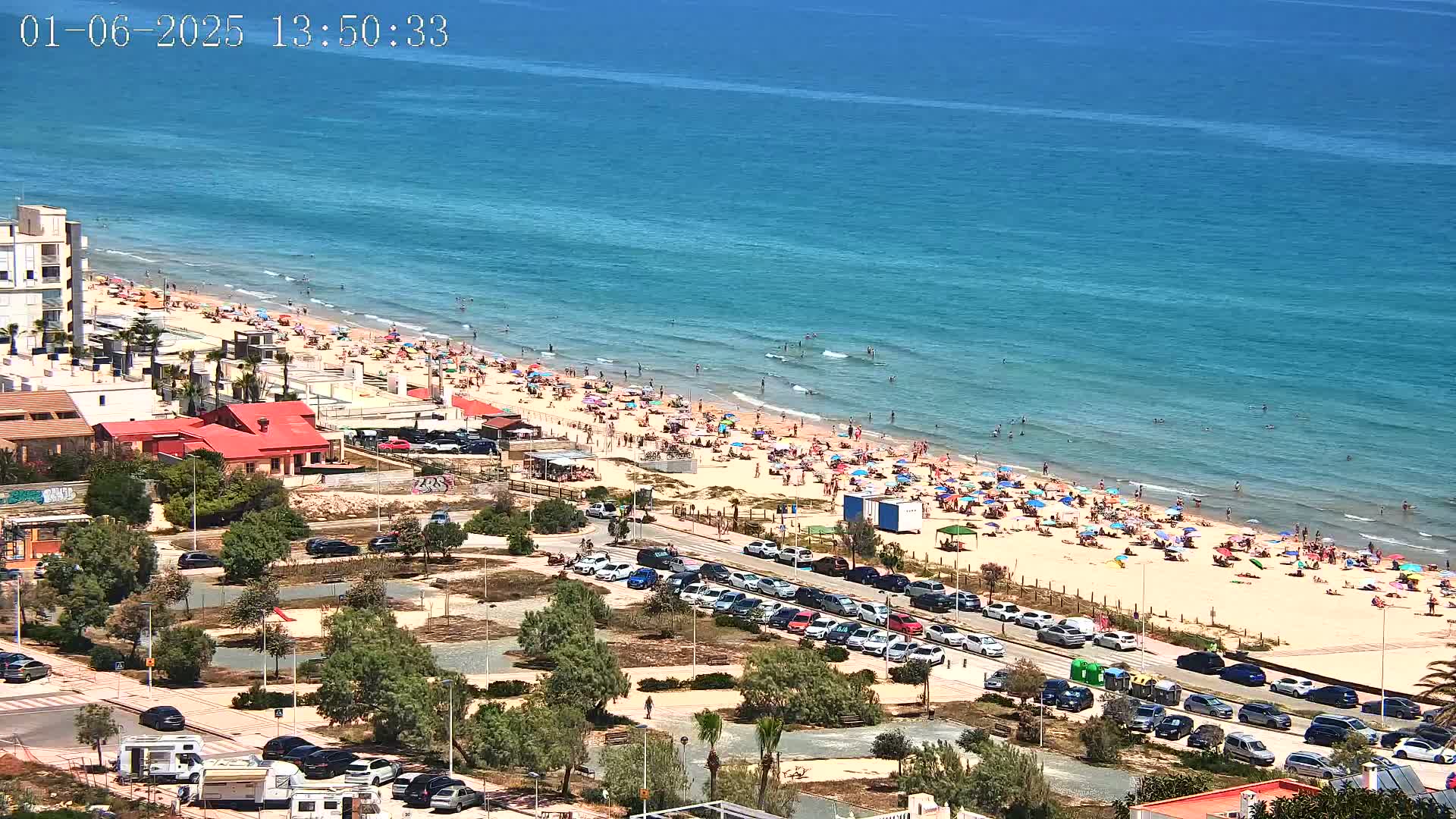 A crowded beach with turquoise water under a sunny sky is viewed from above, showing a parking lot and buildings near the shore.