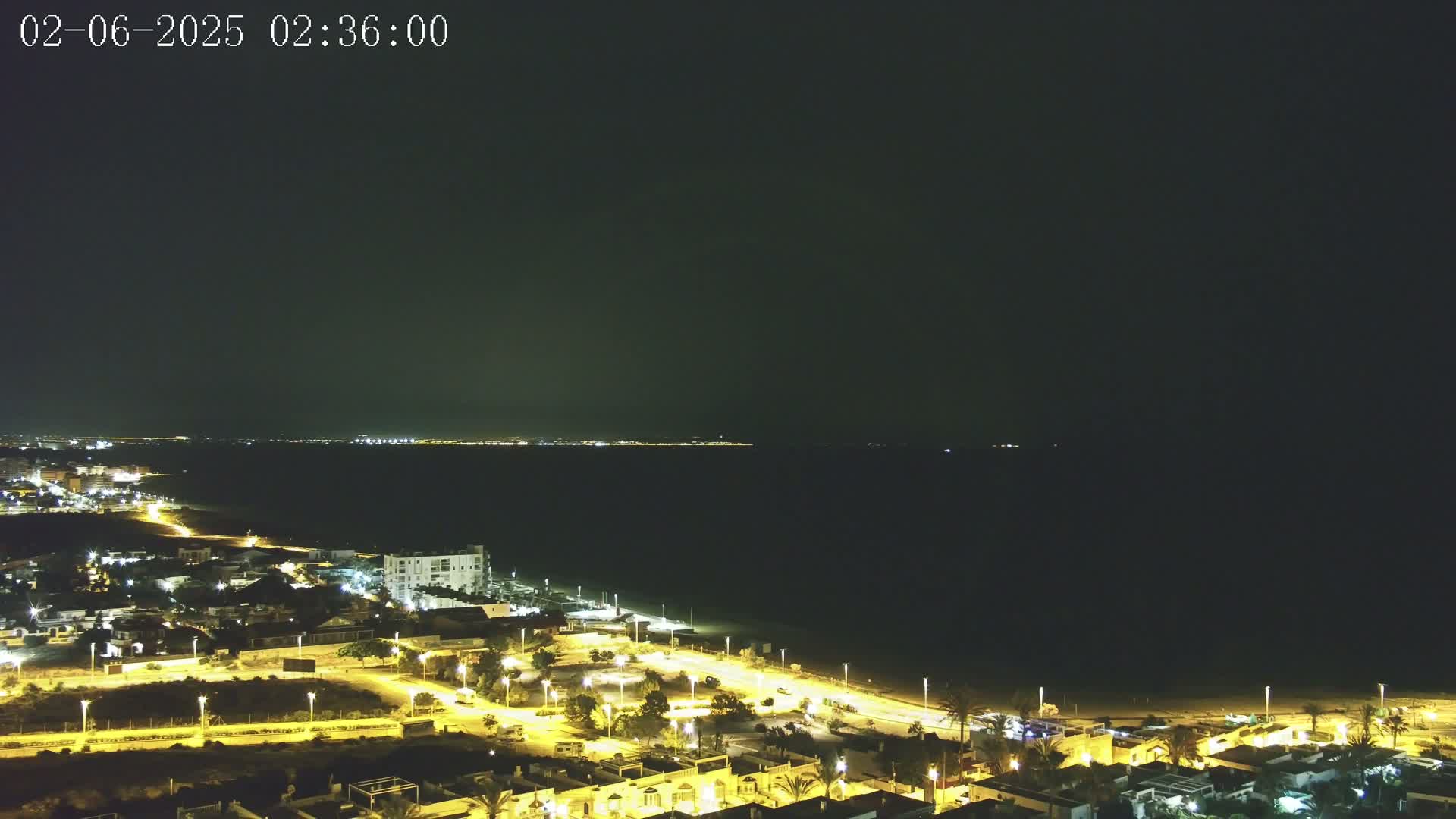 A nighttime coastal scene shows a dark ocean with a faintly lit distant shoreline, and a closer, brightly lit town with streets and buildings extending to a beach.