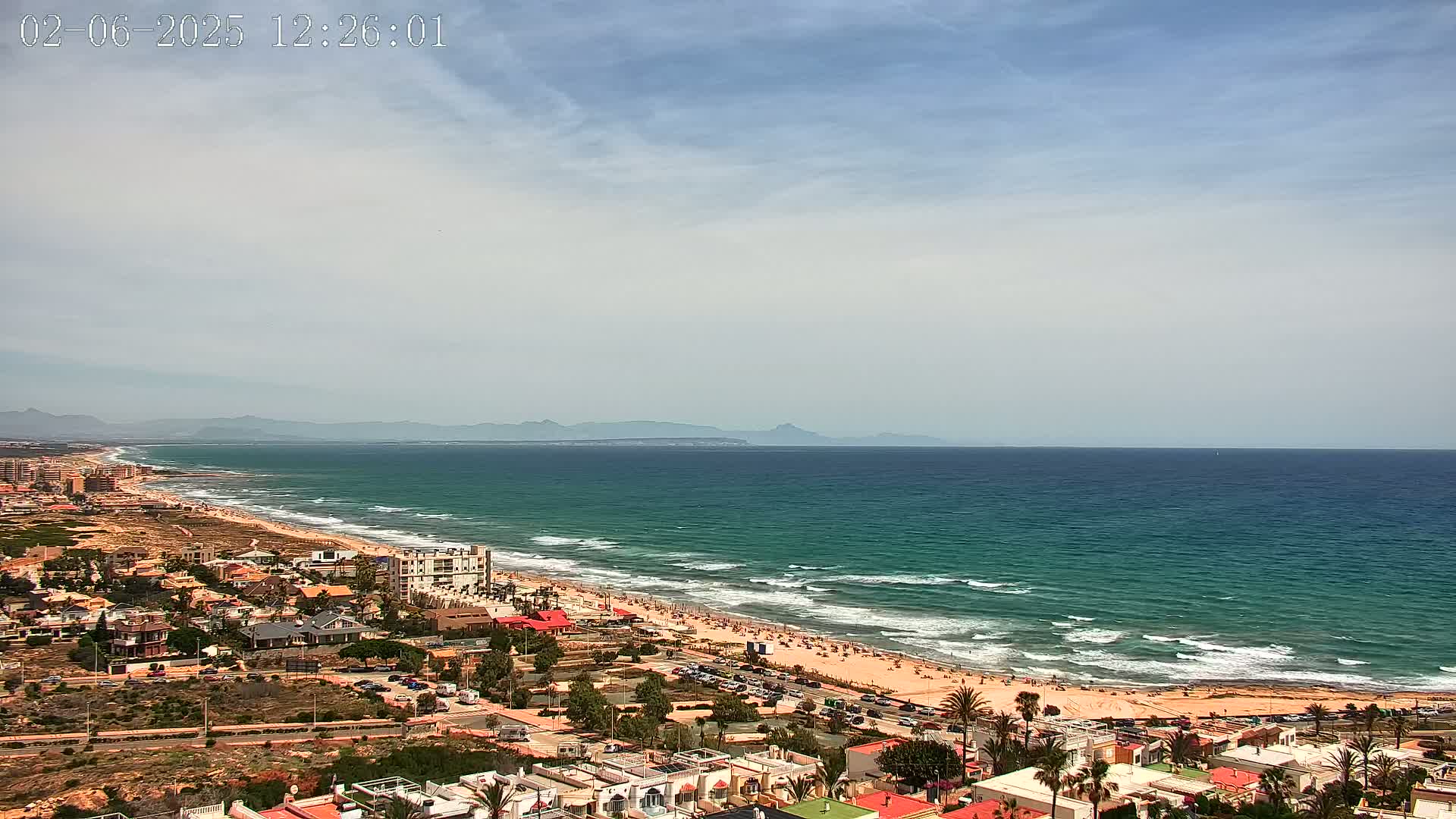 A partly cloudy day reveals a long sandy beach with light waves lapping the shore, backed by a line of low buildings and extending to distant mountains.