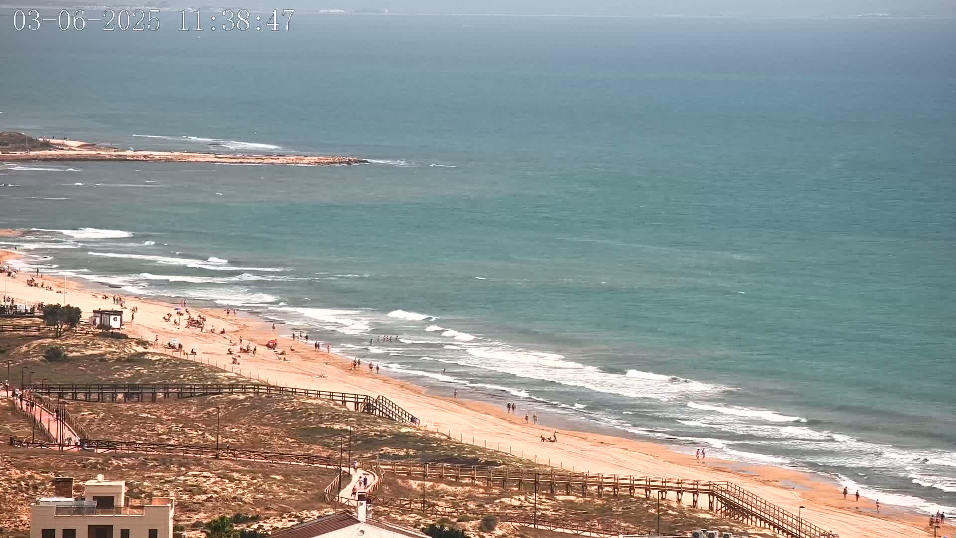 A sandy beach with scattered people is bordered by wooden walkways and overlooks a teal ocean with small waves under a partly cloudy sky.
