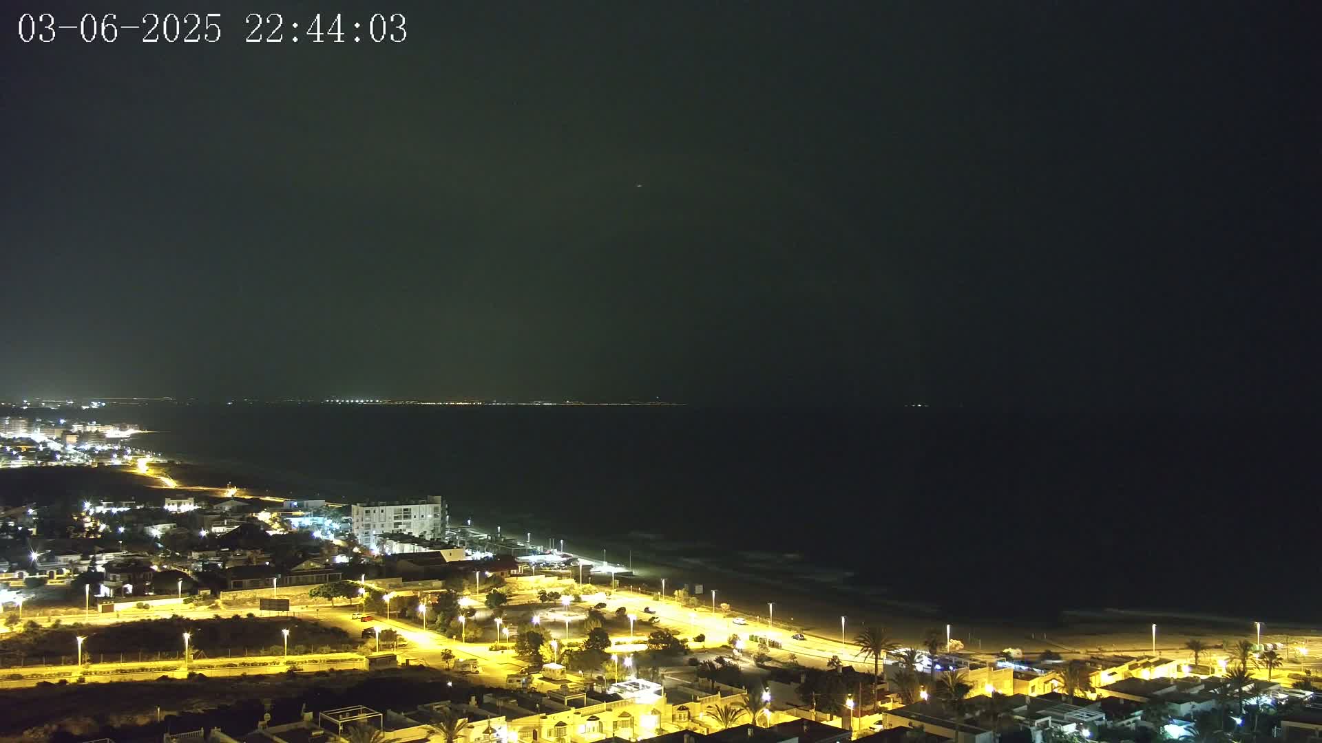 A nighttime coastal scene shows a dark ocean, a beach with illuminated buildings and streets along the shore under a clear, dark sky.