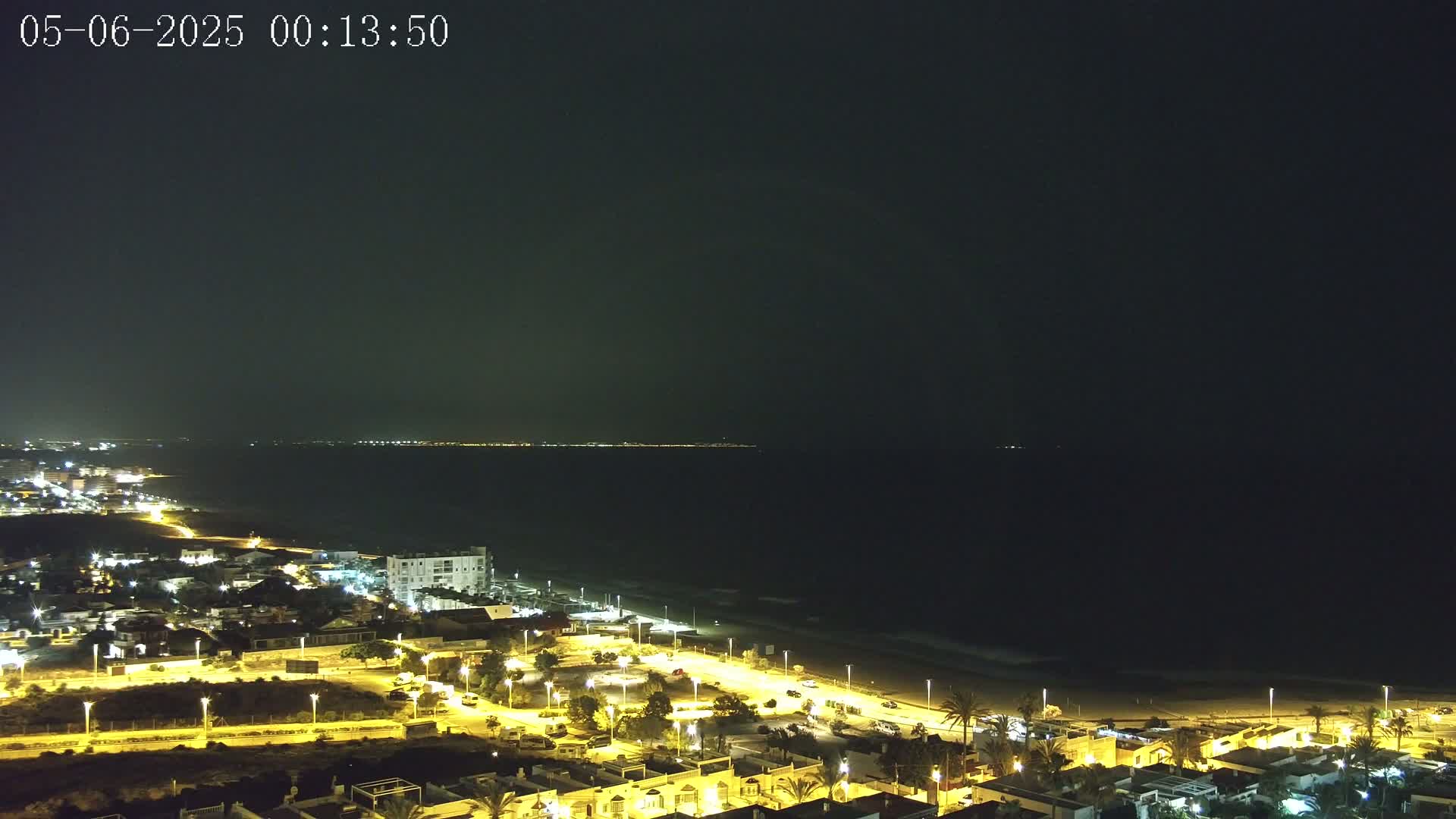 A nighttime coastal town illuminated by streetlights stretches along a dark ocean under a clear, dark sky.
