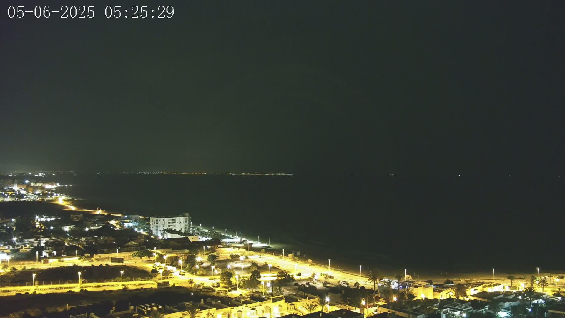 A nighttime, coastal town with illuminated streets and buildings is adjacent to a dark ocean under a clear, dark sky.