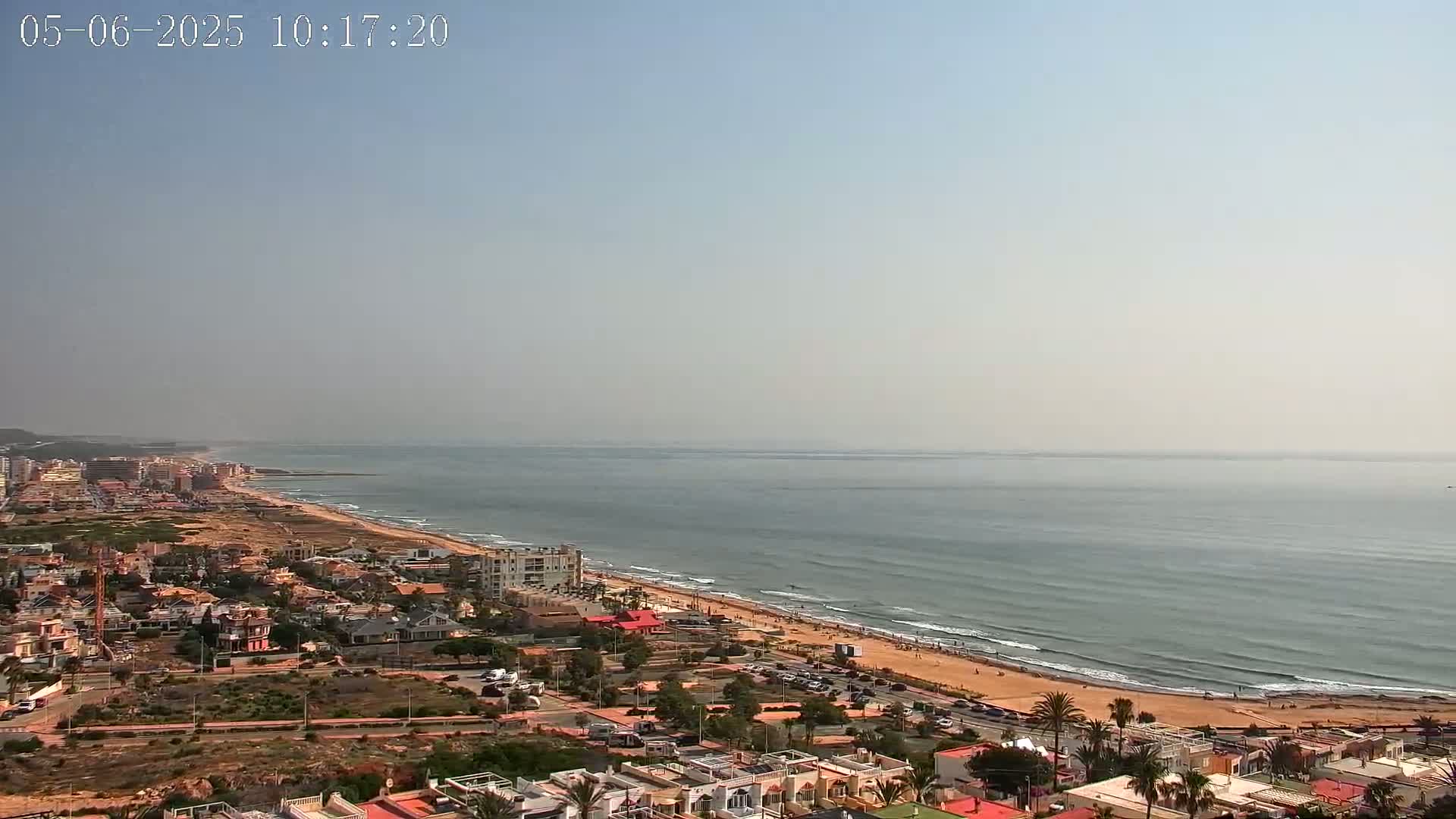 A hazy, sunny day reveals a coastal town overlooking a calm ocean with a sandy beach and gentle waves.