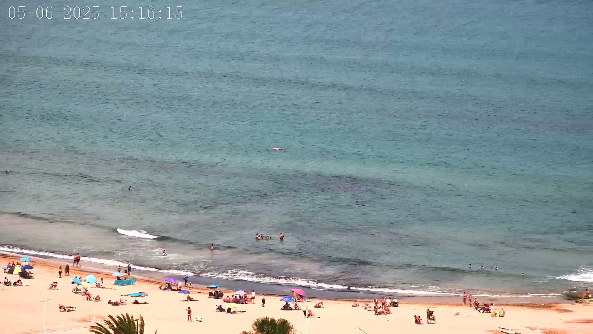 A sandy beach with scattered sunbathers and umbrellas borders a calm, teal ocean with a few people swimming.