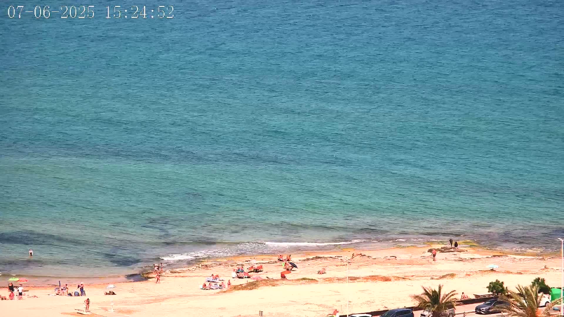 A sandy beach with scattered sunbathers is adjacent to a calm, teal-colored ocean under a sunny sky.