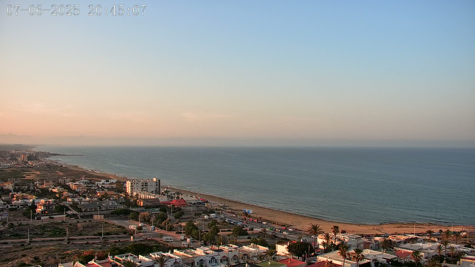An aerial view shows a coastal town at sunset, with a calm ocean extending to a hazy horizon.
