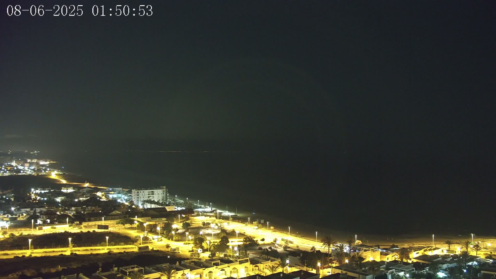 A nighttime coastal town brightly lit, with a dark sea stretching to a dark horizon under a clear sky.