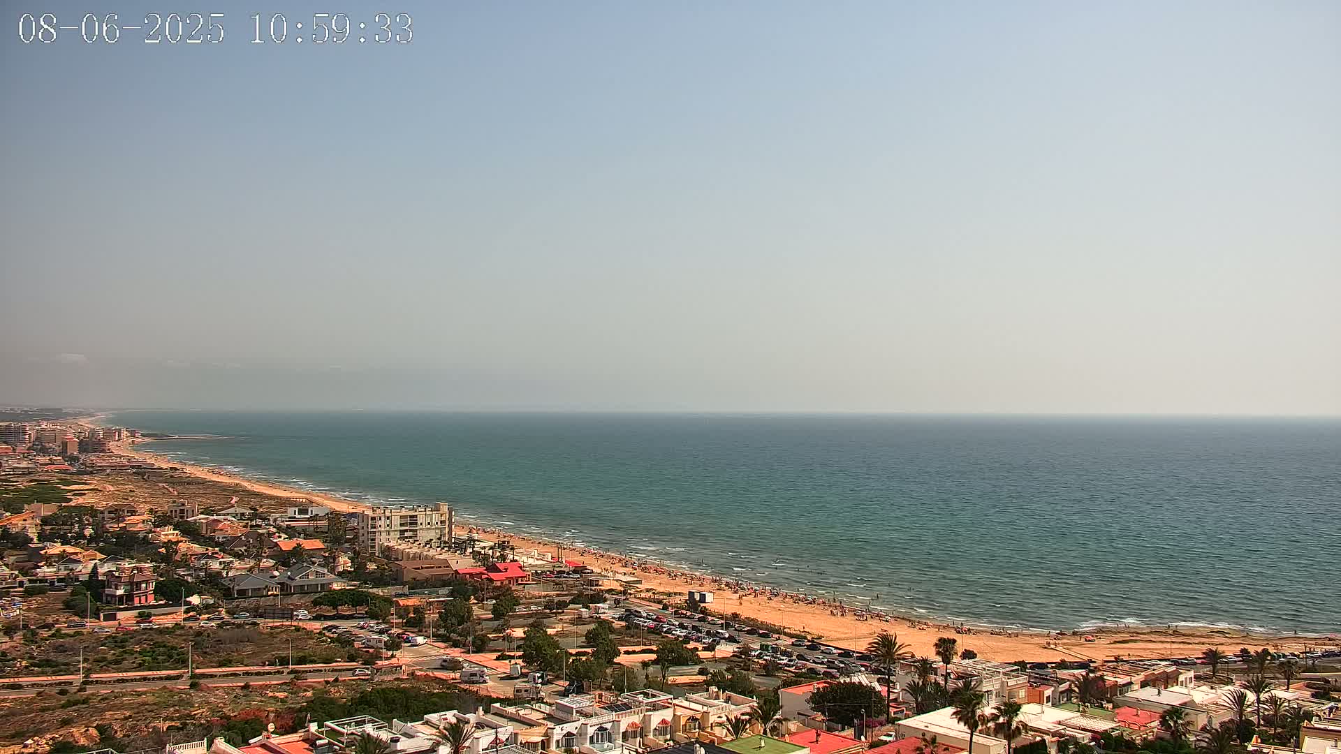 An aerial view shows a long sandy beach with people on it, next to a town of low-rise buildings, all bordering a calm, teal ocean under a hazy sky.