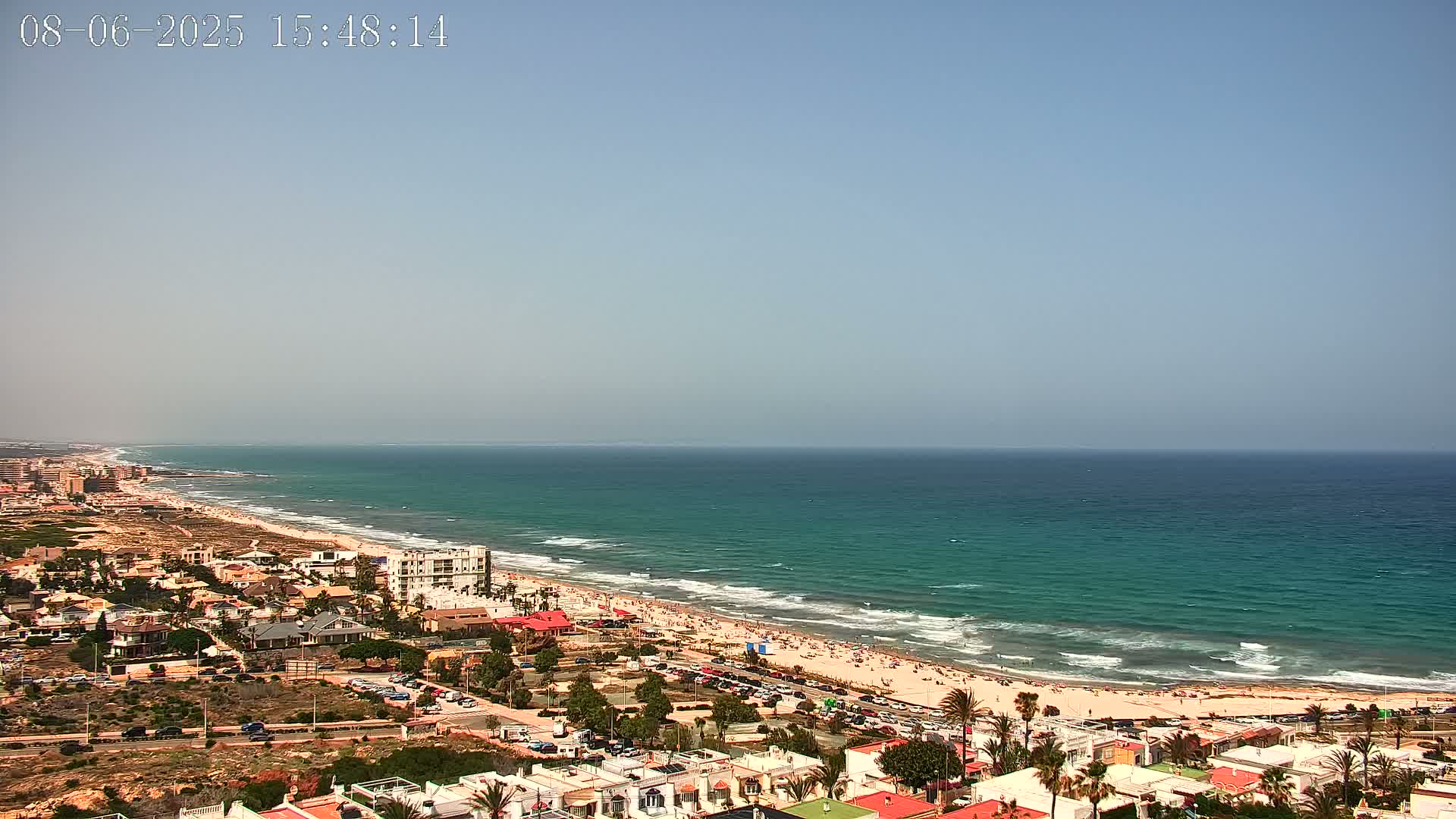 A beach with turquoise water and small waves is seen from an elevated vantage point, showing a town with houses and parked cars extending inland under a clear, bright sky.