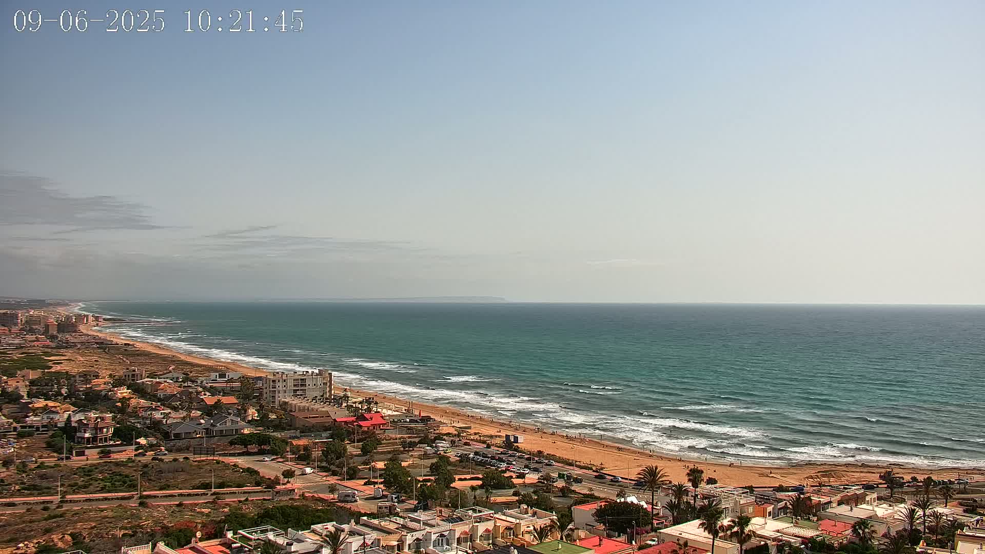 A coastal town overlooks a long sandy beach and a teal ocean with small waves under a mostly clear, slightly hazy sky.
