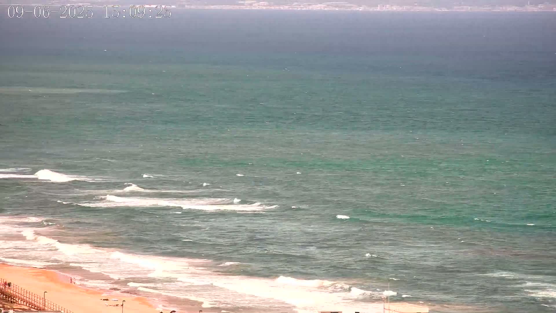 A sandy beach with small waves and a calm, teal-colored sea stretches to a distant shoreline under an overcast sky.